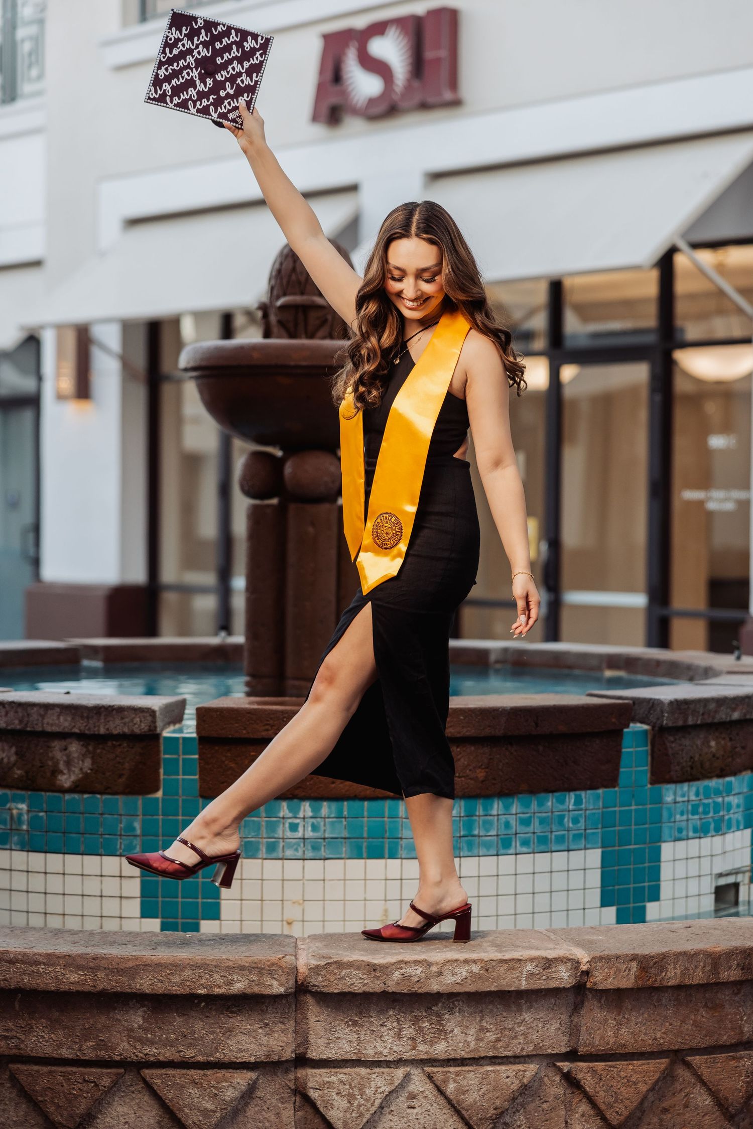 Graduate in black dress and yellow sash poses playfully by turquoise-tiled fountain during graduation photoshoot.