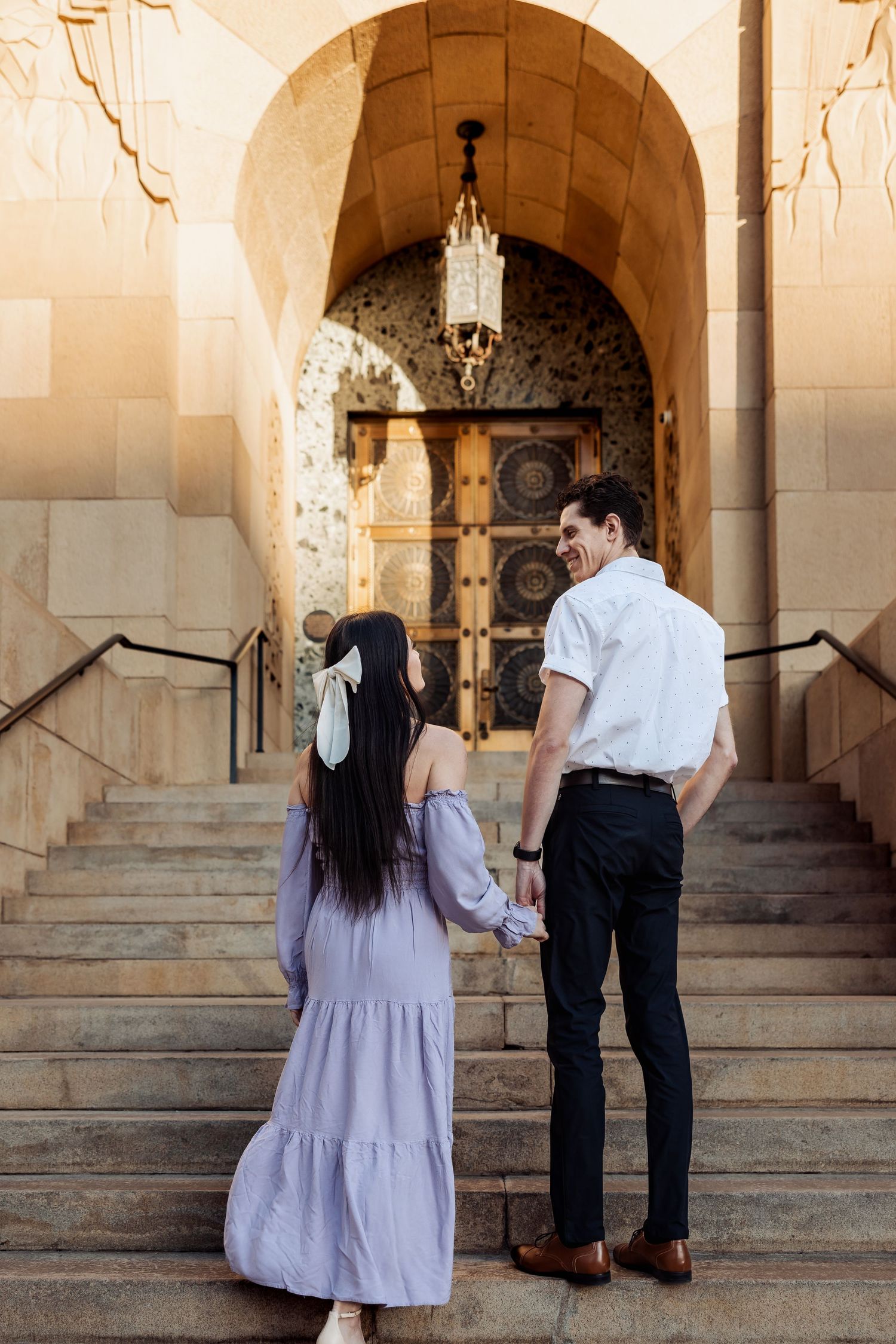 A couple stands on stone steps leading to an arched entryway with a hanging vintage lantern in warm lighting.