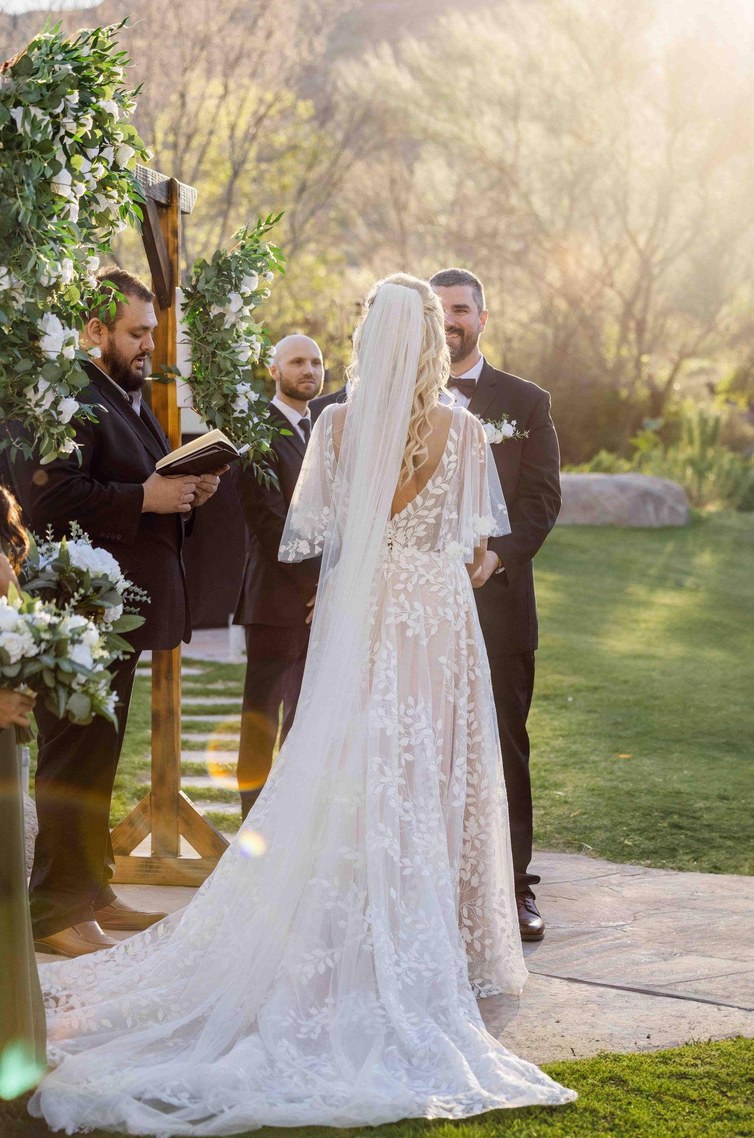 Outdoor wedding ceremony at sunset with a decorative floral arch and flowing white lace veil in the foreground.