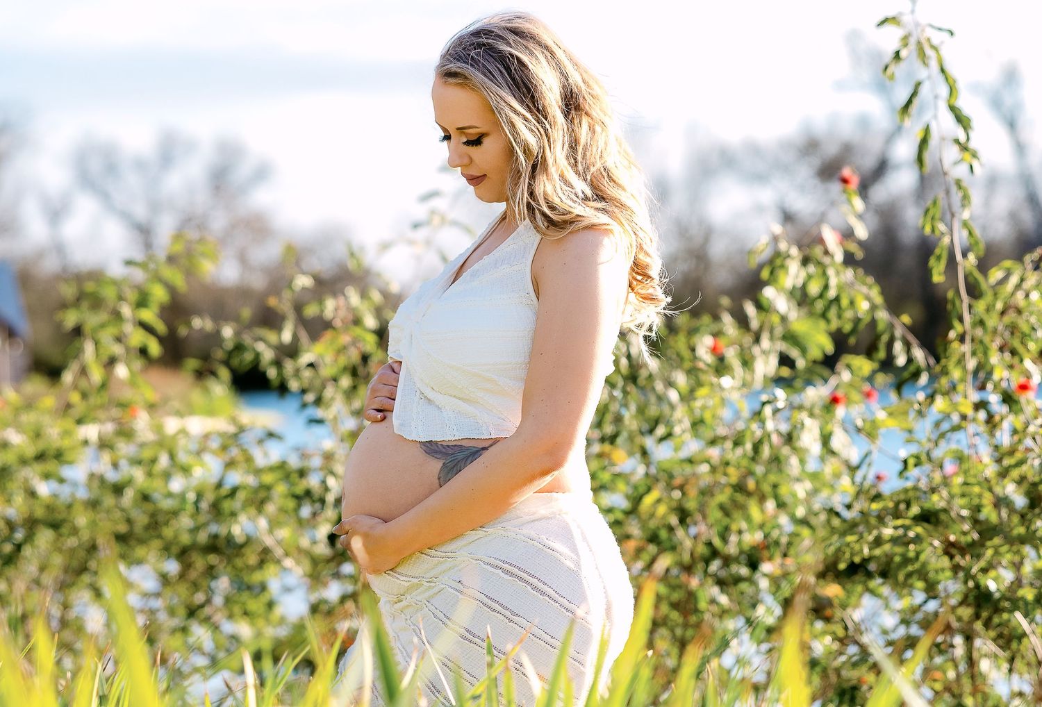 A pregnant woman in a white dress stands in a sunny garden with blooming flowers and greenery.