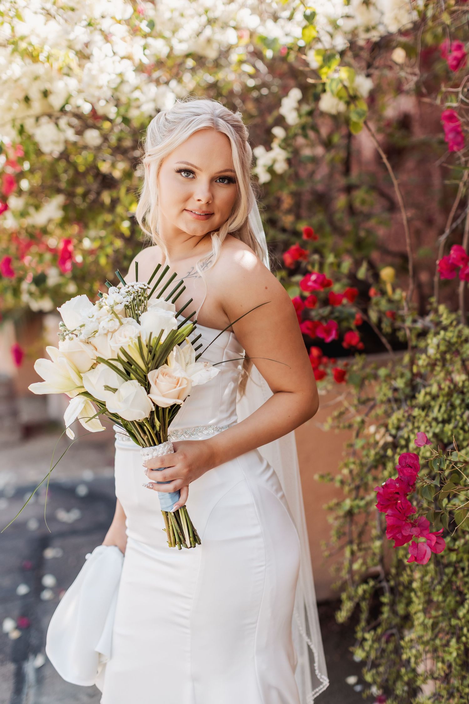 A bride in a white strapless wedding gown holds a bouquet of white lilies against a backdrop of pink bougainvillea flowers.