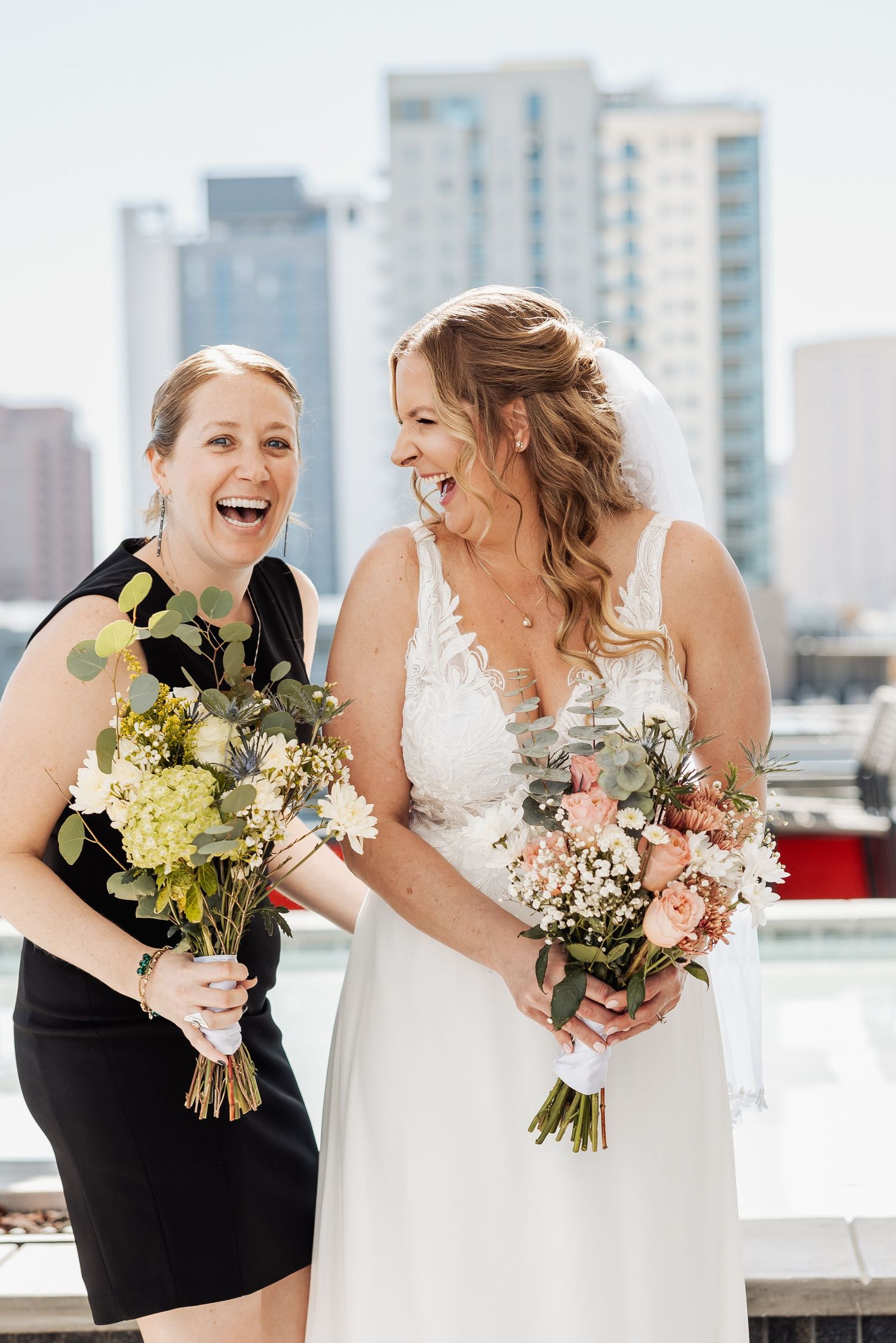 Wedding party members in black and white dresses share joyful laughter while holding floral bouquets on a rooftop.