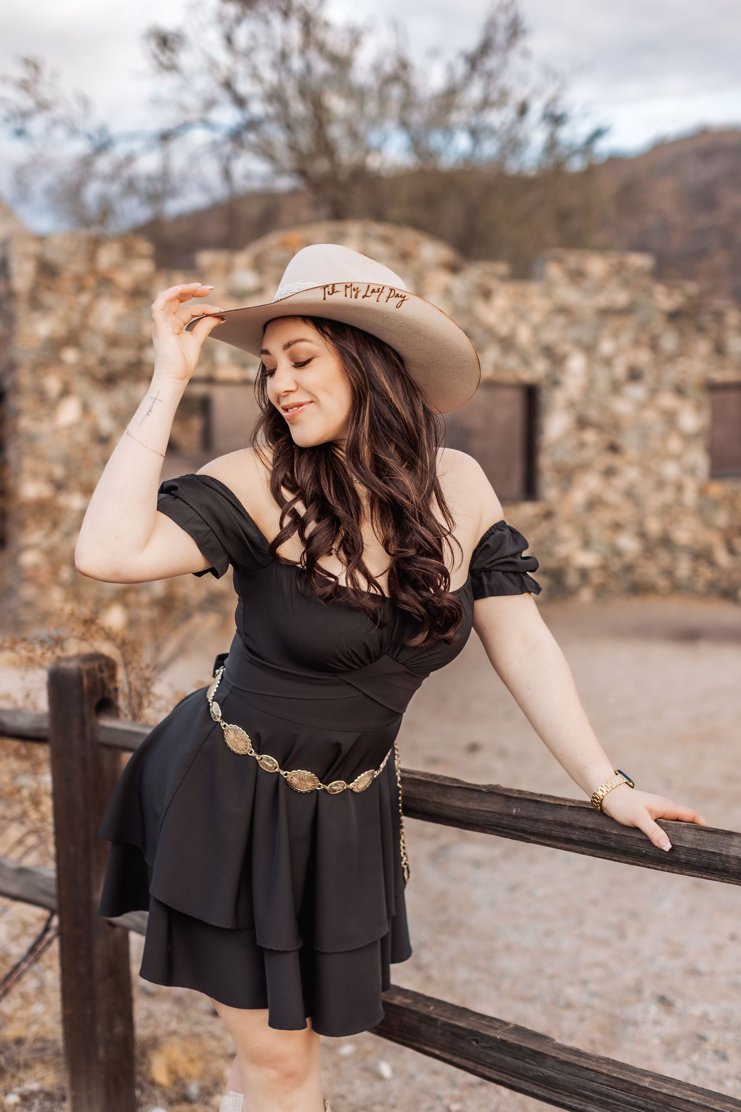 Western style outfit with black dress, concho belt, and cowboy hat against stone wall and wooden fence in desert setting.