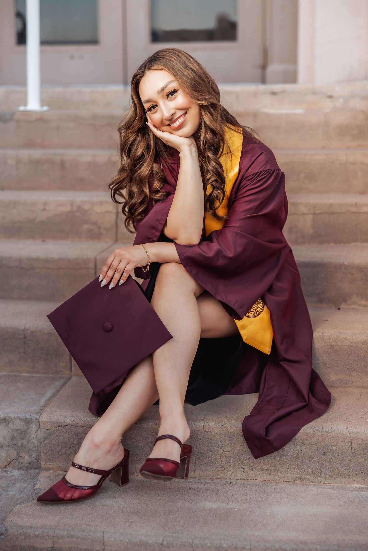 Graduate in burgundy gown poses on stone steps while wearing yellow blouse and burgundy shoes for graduation photos.