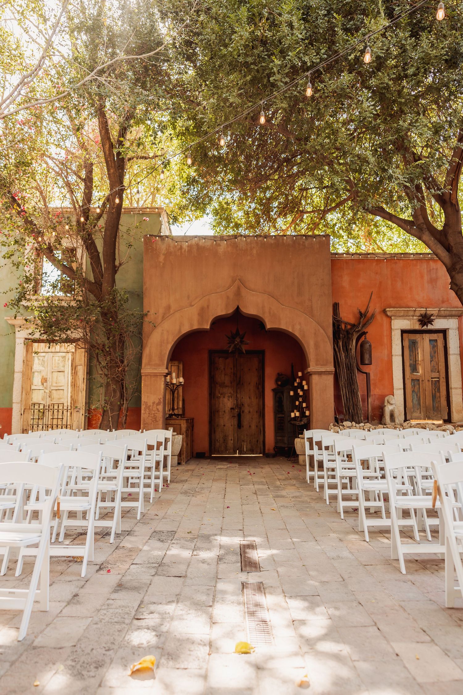Rustic outdoor wedding venue with white chairs set up for ceremony under trees with terra cotta walls.