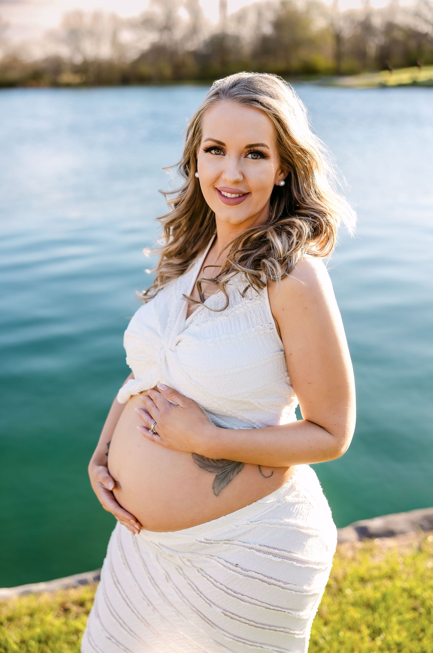 Outdoor pregnancy photography session near water with white dress and natural lighting.