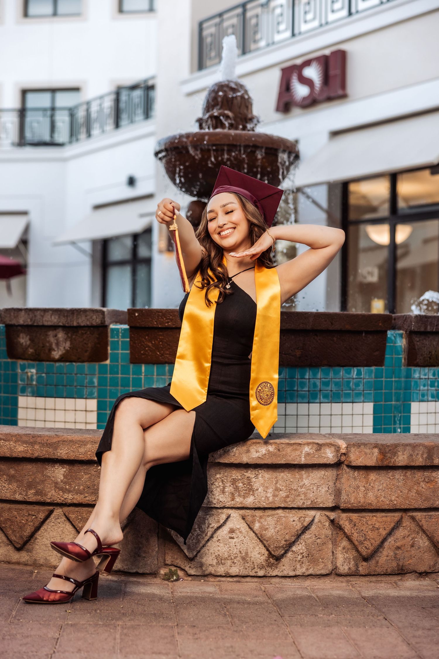 Joyful graduate in black dress and yellow stole laughs while sitting on brick wall near campus building.