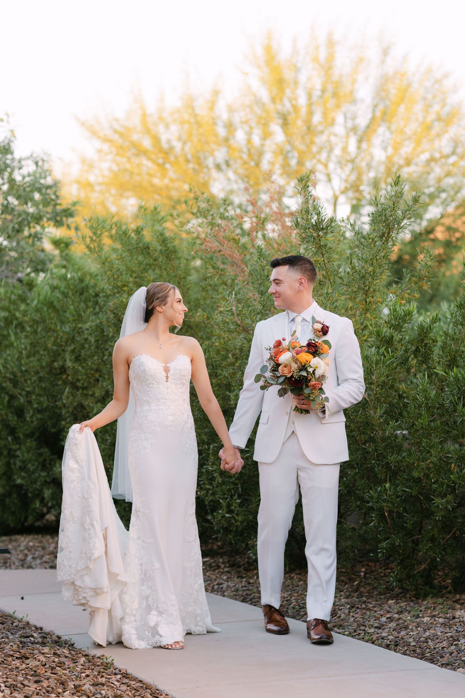 Newlyweds walking together on path with desert landscaping and autumn foliage.