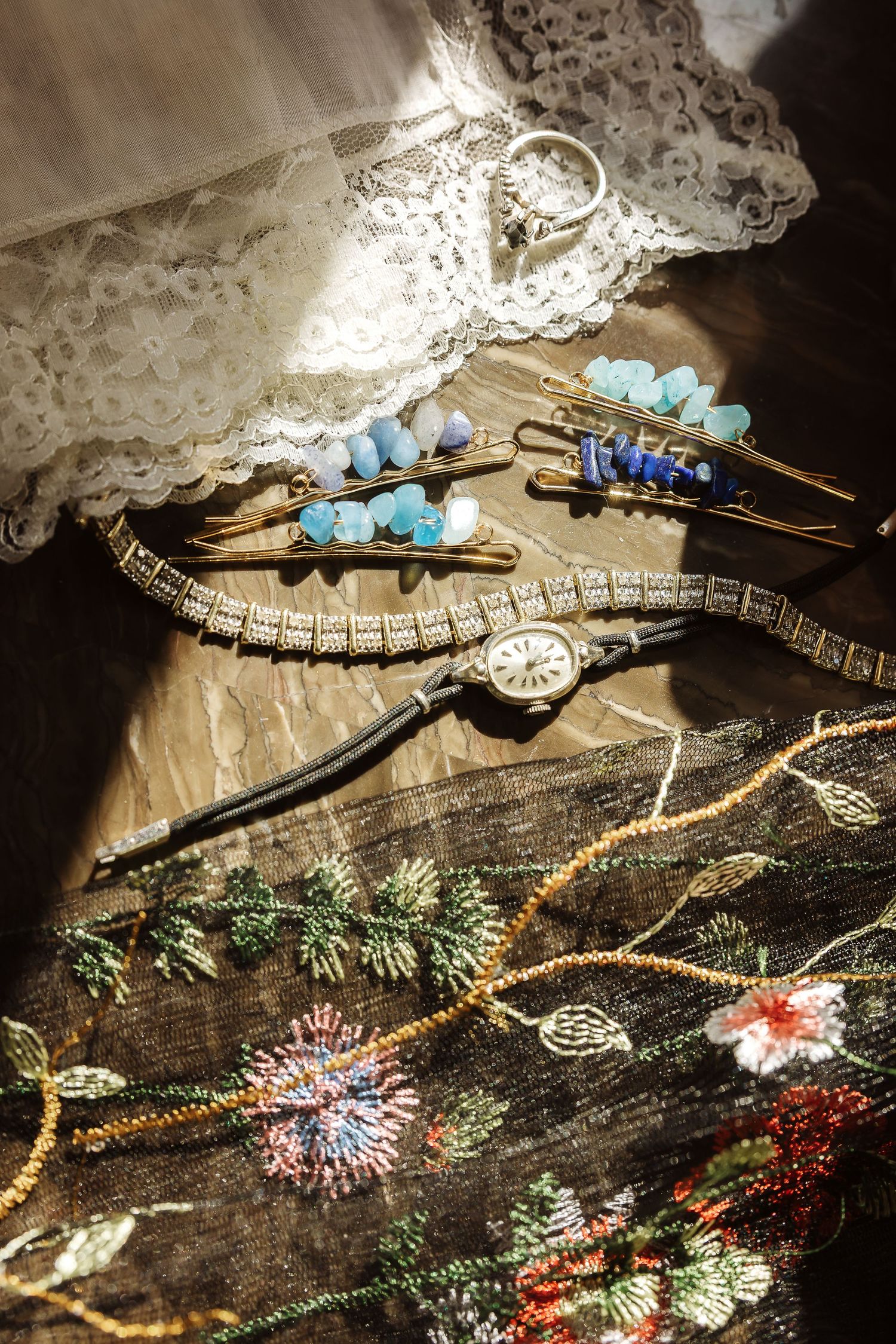 Wedding accessories including lace, blue floral hair clips, a vintage watch and embroidered fabric in soft lighting.