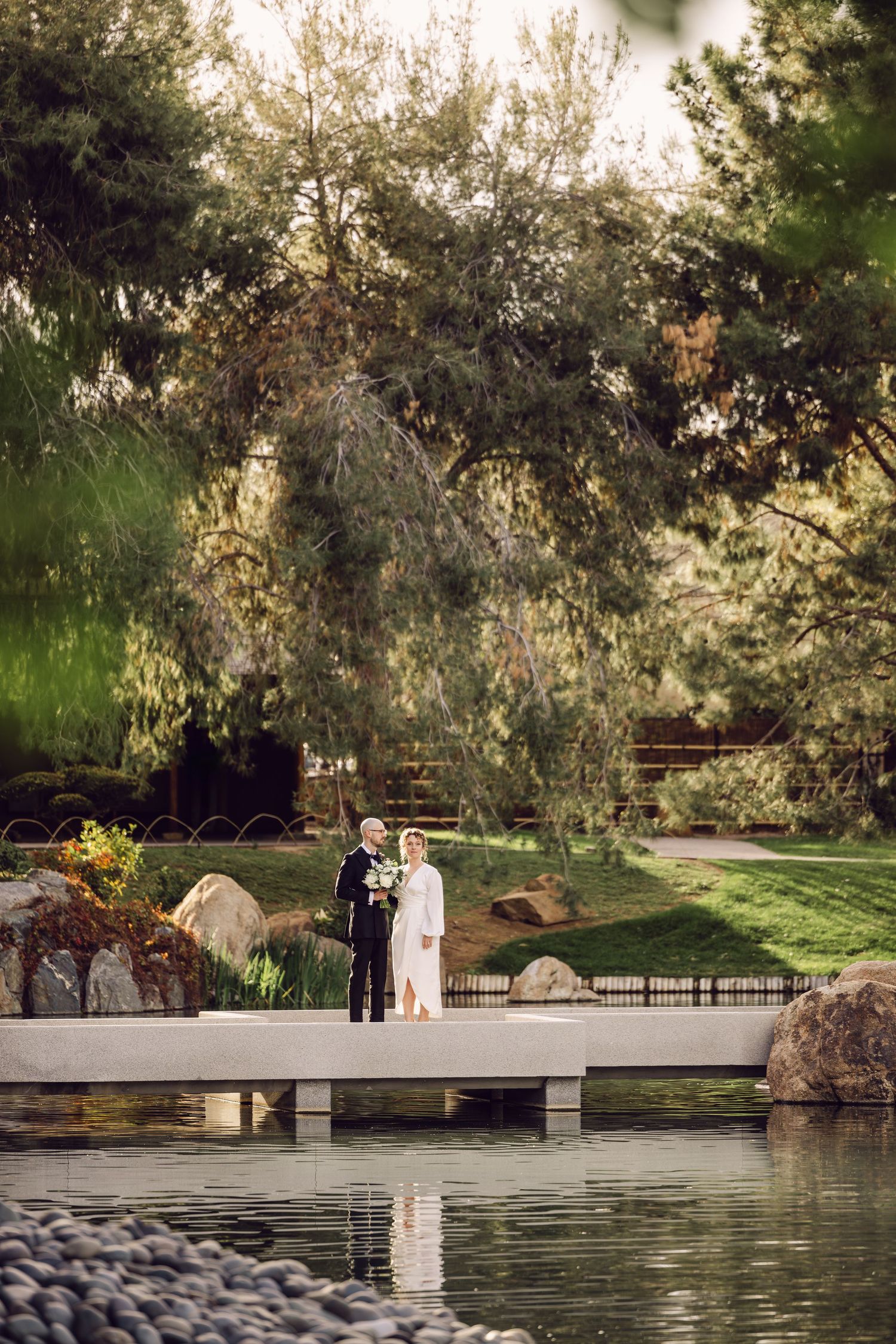 A couple stands on a white bridge overlooking a serene pond in a lush garden setting during a romantic moment.