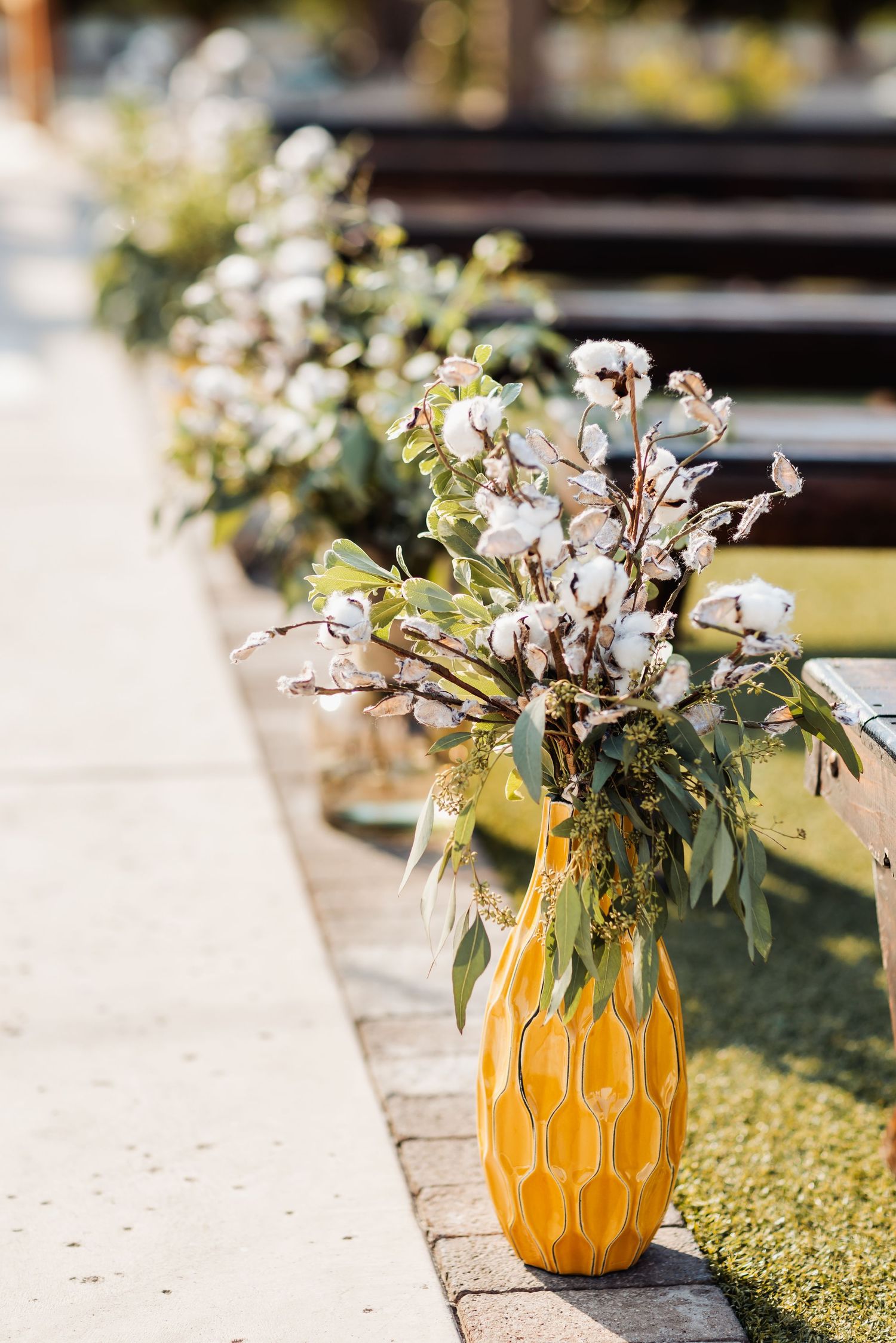 Yellow ceramic vase filled with cotton stems and greenery as wedding aisle decoration.