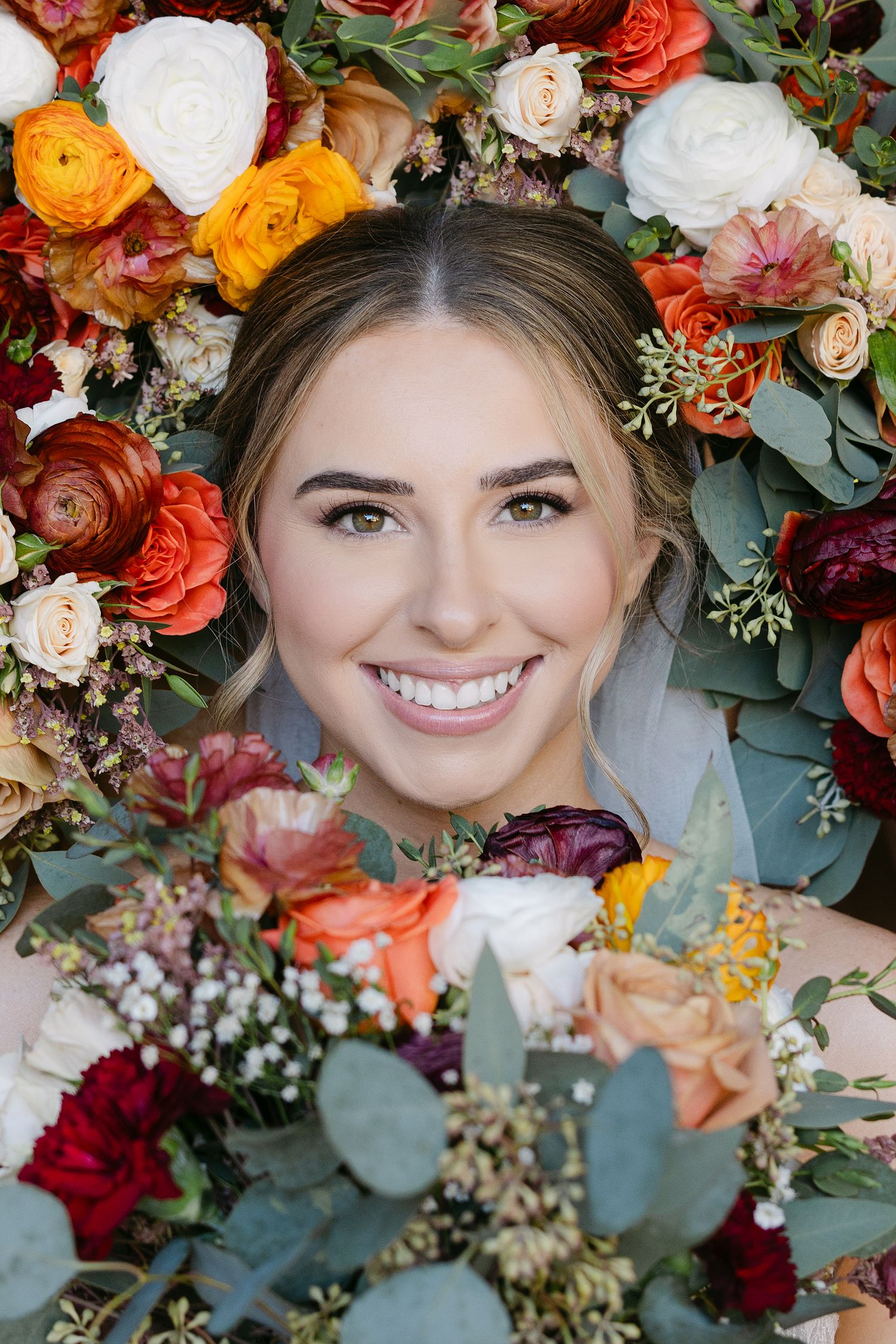 Beautiful floral arrangement with orange, white and burgundy blooms framing a smiling bride against a blue background.