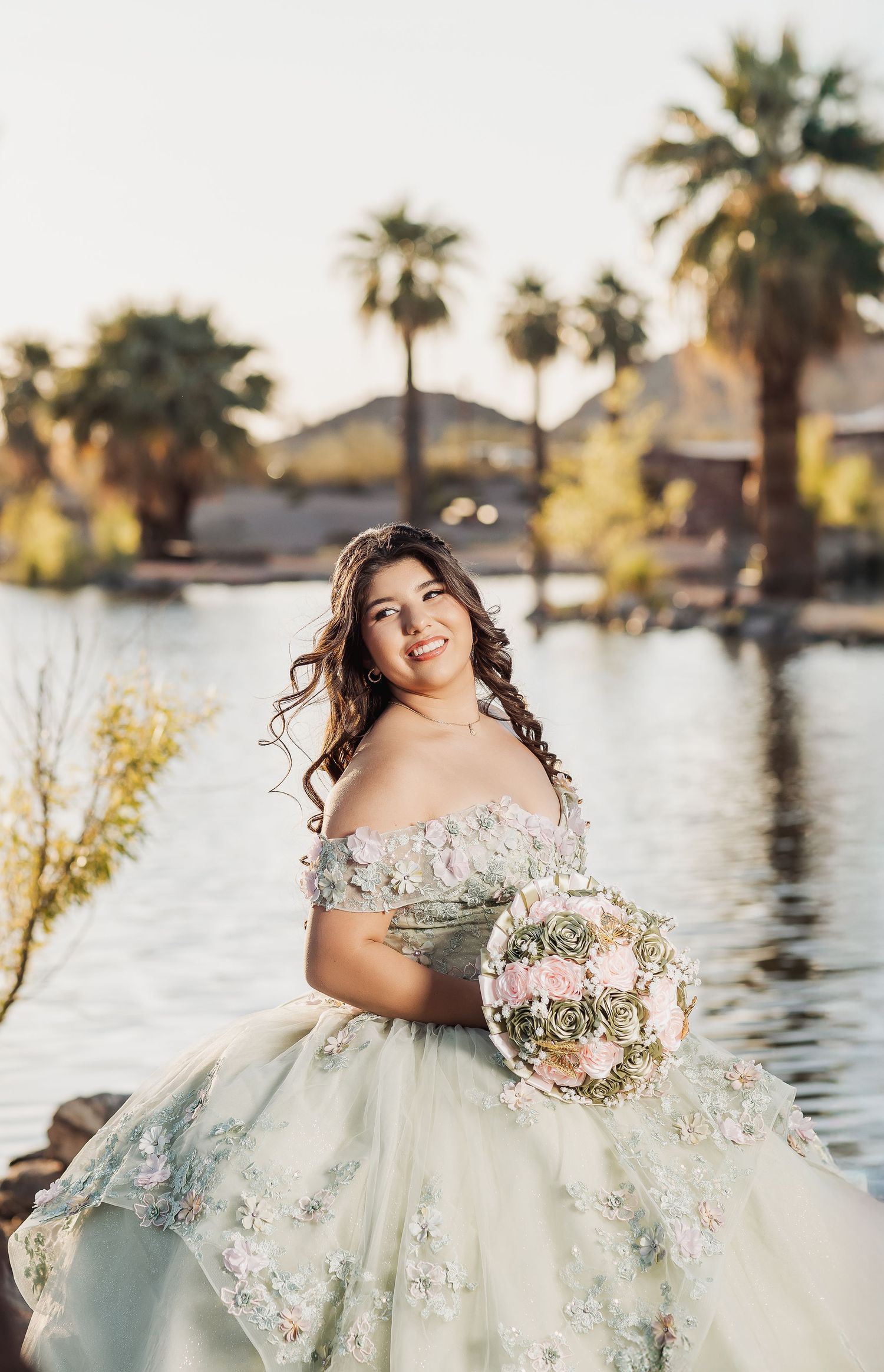 A bride in an ethereal white gown with floral details poses gracefully by a lakeside during golden hour.