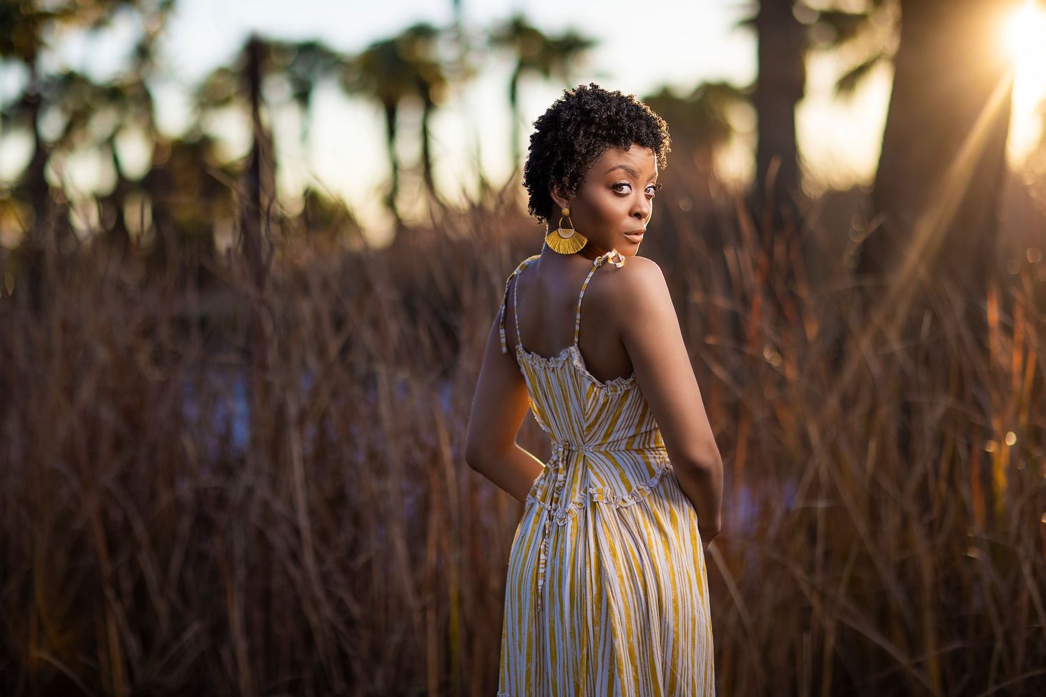 Someone in an elegant striped dress poses in tall grass at sunset with palm trees silhouetted in the background.