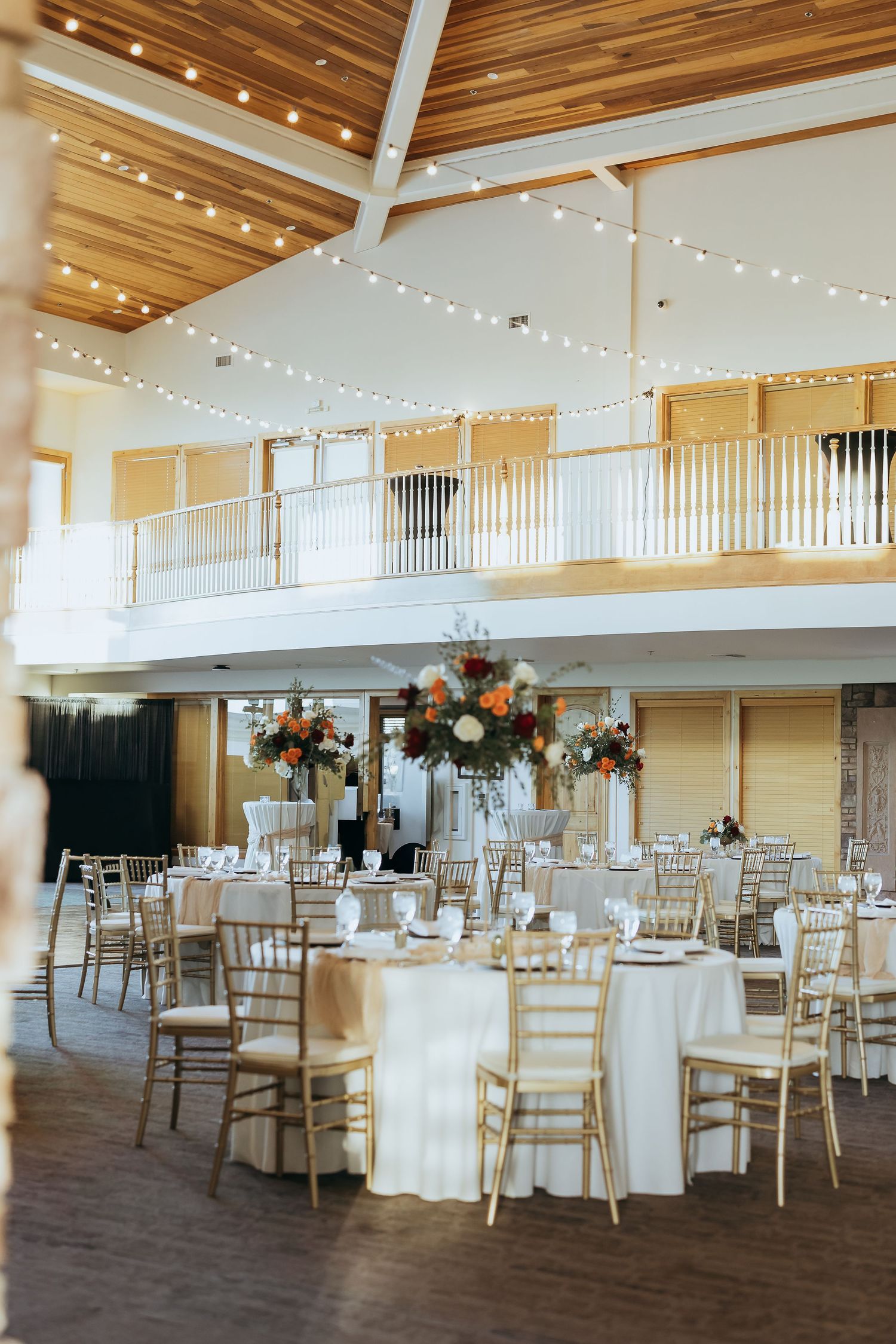 Elegant wedding reception venue with wooden ceiling, string lights, and round tables set with white linens and gold chairs.