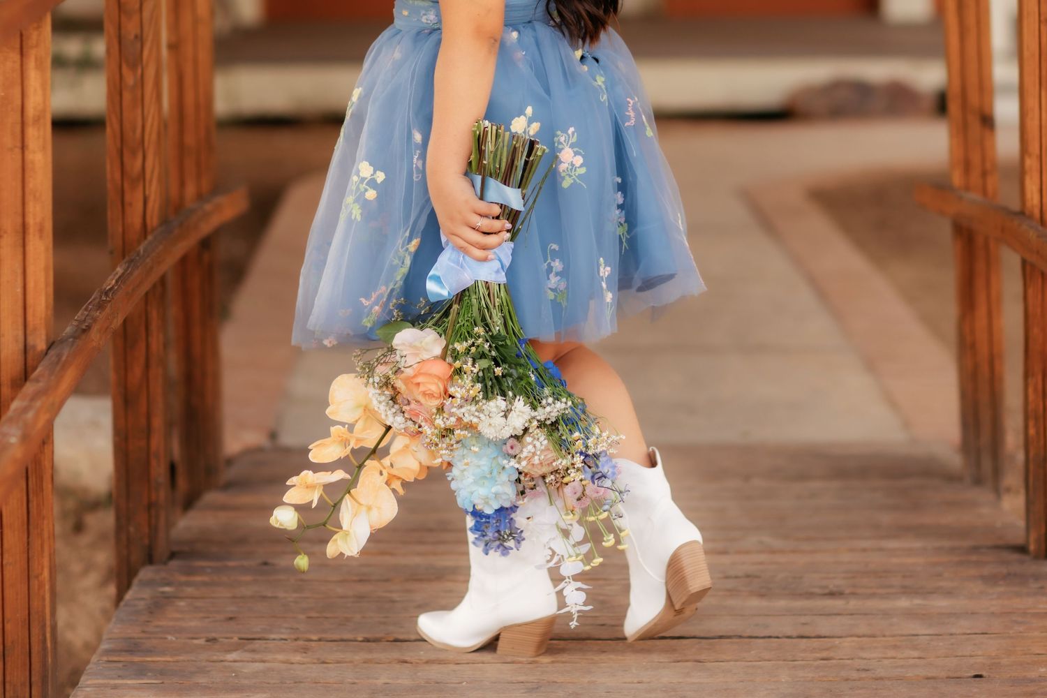 Person in blue tulle dress and white boots stands on wooden bridge holding cascading bouquet of pastel flowers.