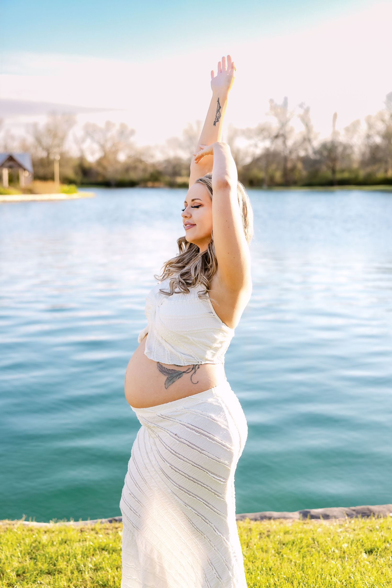 Maternity photoshoot by a lake with white fabric draping and golden sunlight on grassy shore.