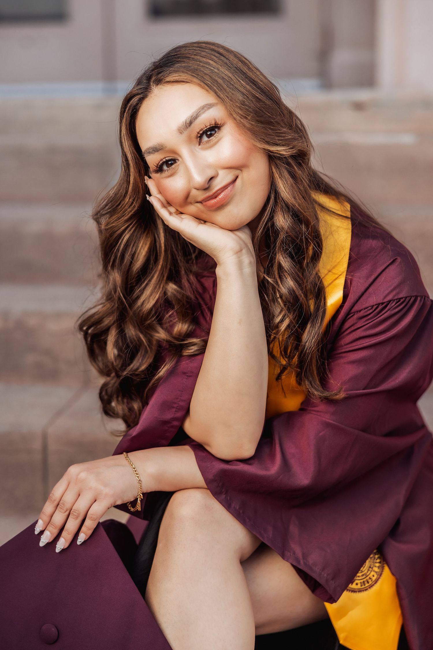 Graduate in maroon gown with gold sash sits on stone steps during graduation photoshoot.