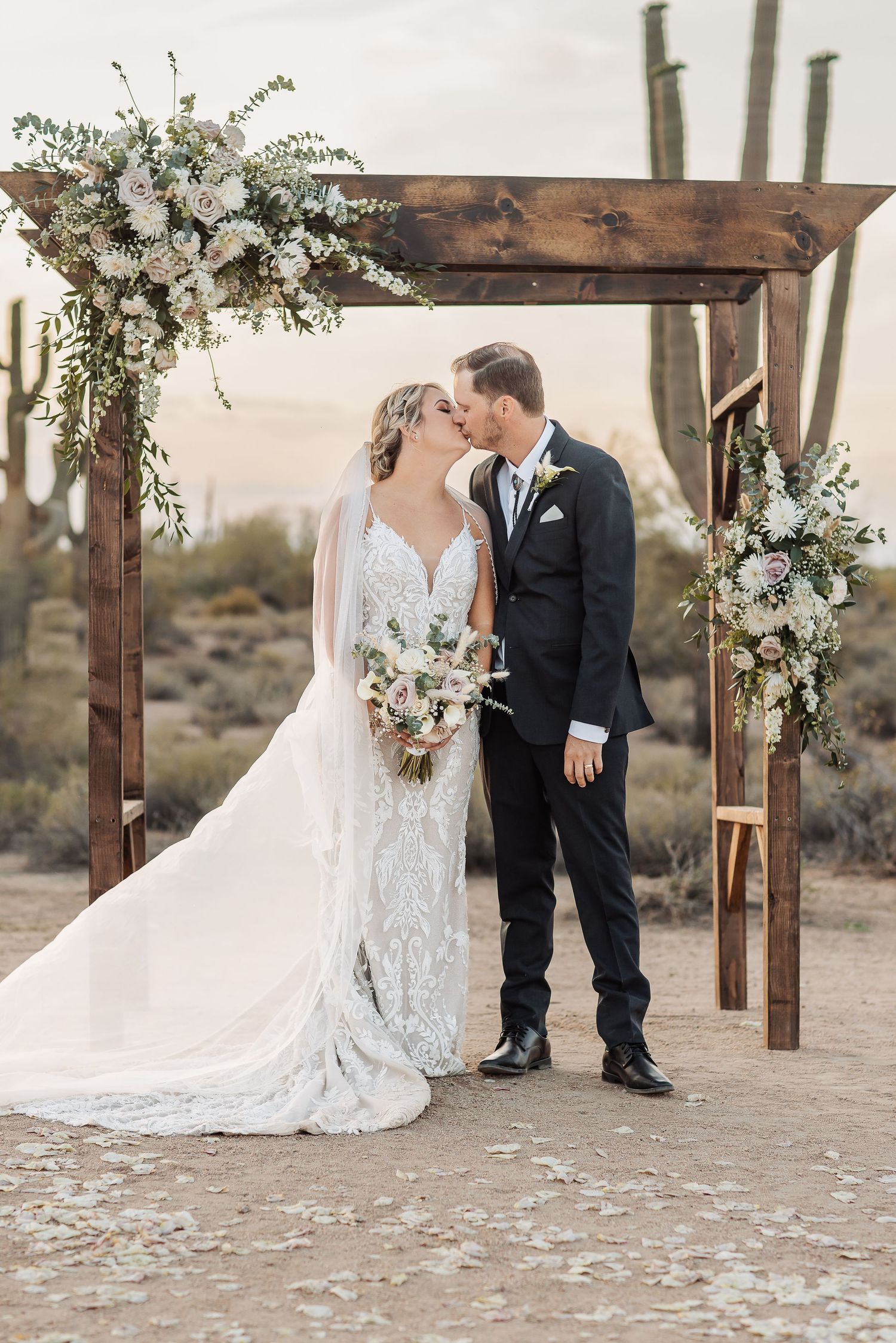 A wedding ceremony under a rustic wooden arch decorated with white floral arrangements in a desert setting.
