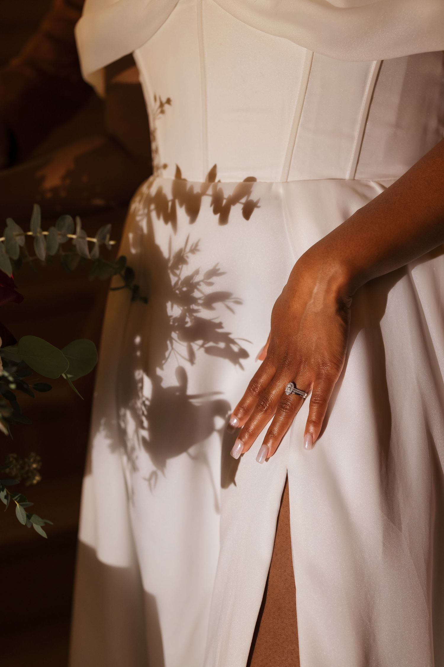 Elegant white dress with a thigh slit and delicate leaf shadows cast across the fabric, with a ring-adorned hand visible.