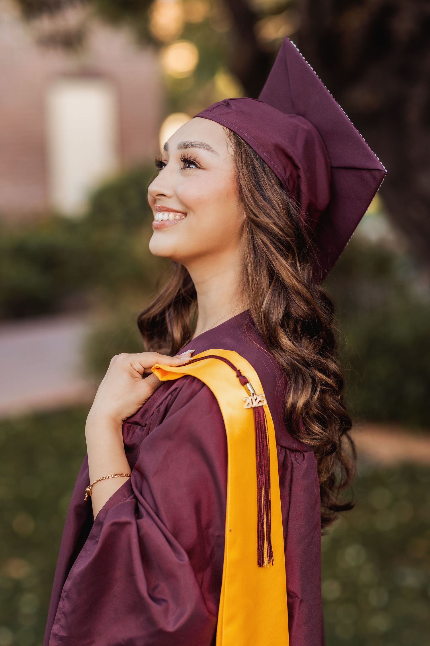 Graduate in maroon cap and gown with long wavy hair poses outdoors with a joyful smile.
