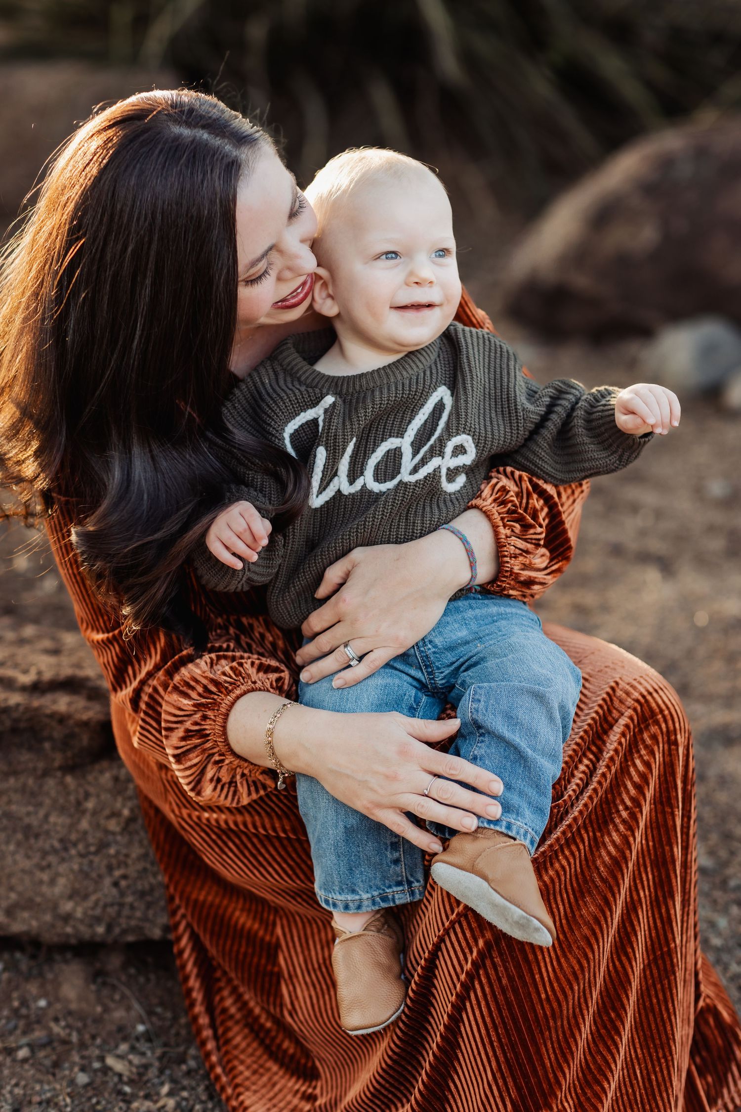 Parent in rust velvet dress snuggles smiling toddler wearing denim and casual dark shirt outdoors in nature.