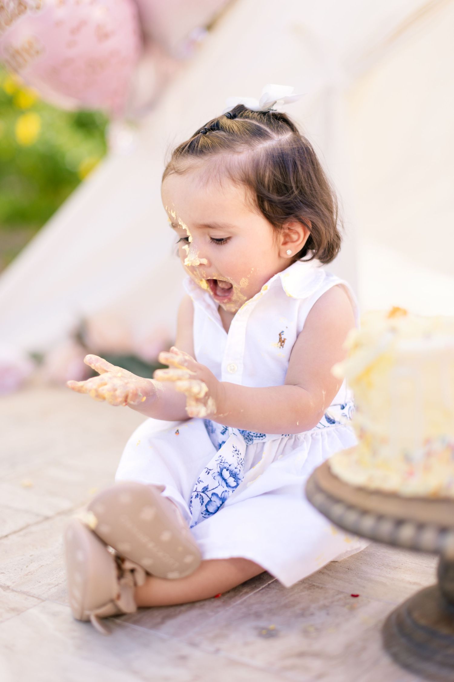 A young child in a white dress sits on the floor and joyfully eats during a birthday celebration.