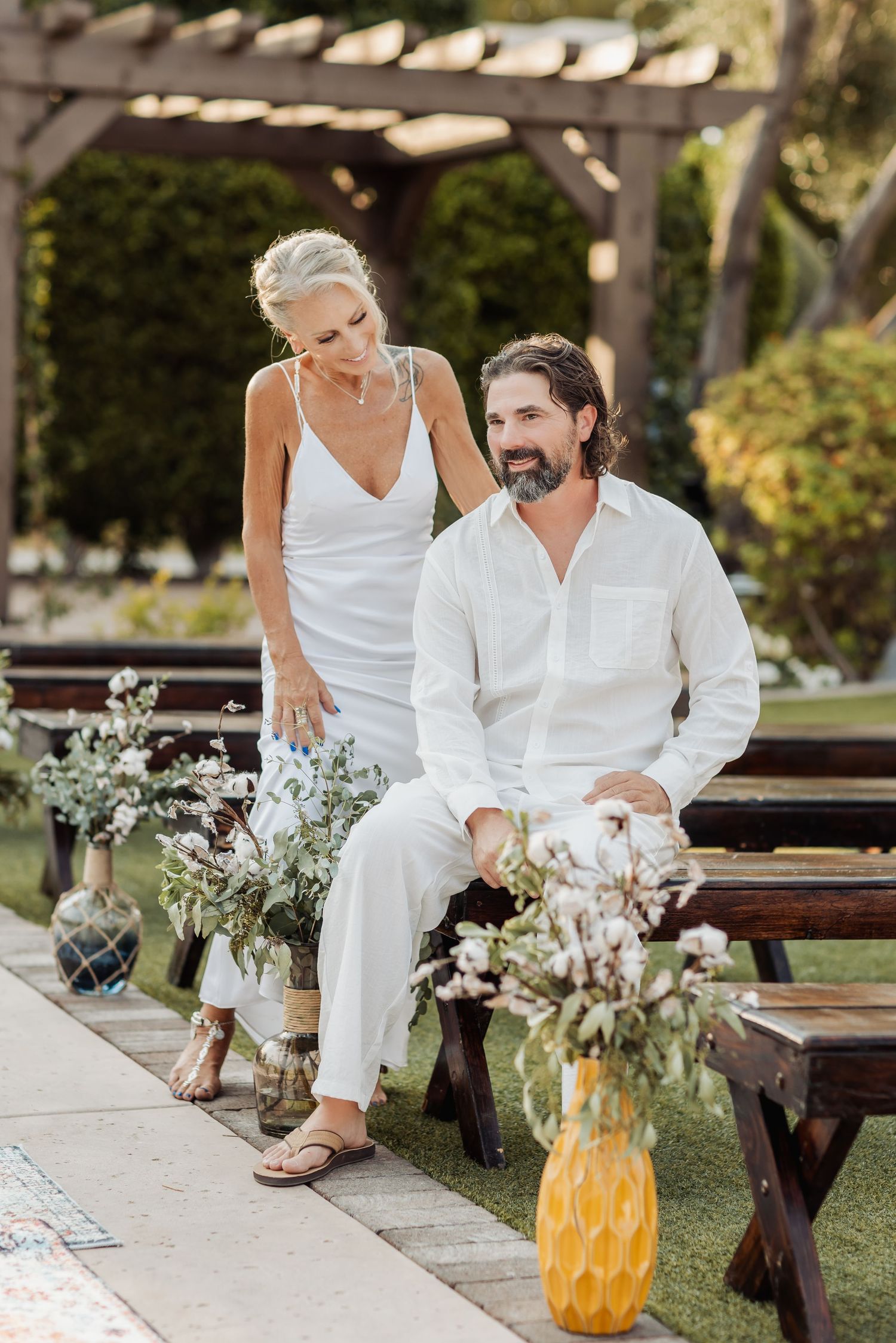Couple in white outfits sharing romantic moments on wooden benches with floral decorations.