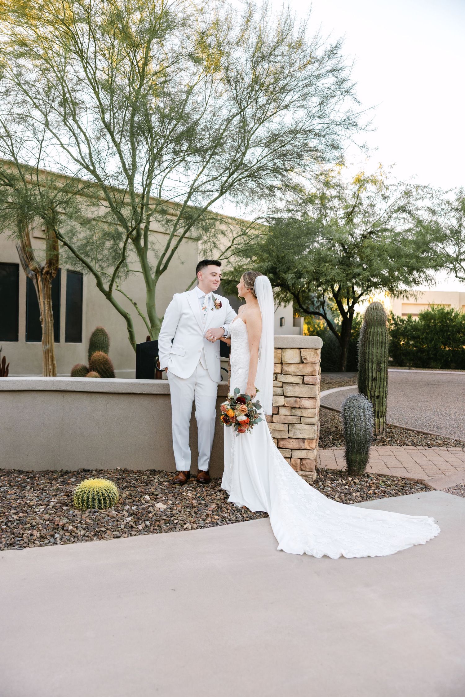 A wedding couple poses for photos in a desert garden setting with white stone walls and cacti in the background.
