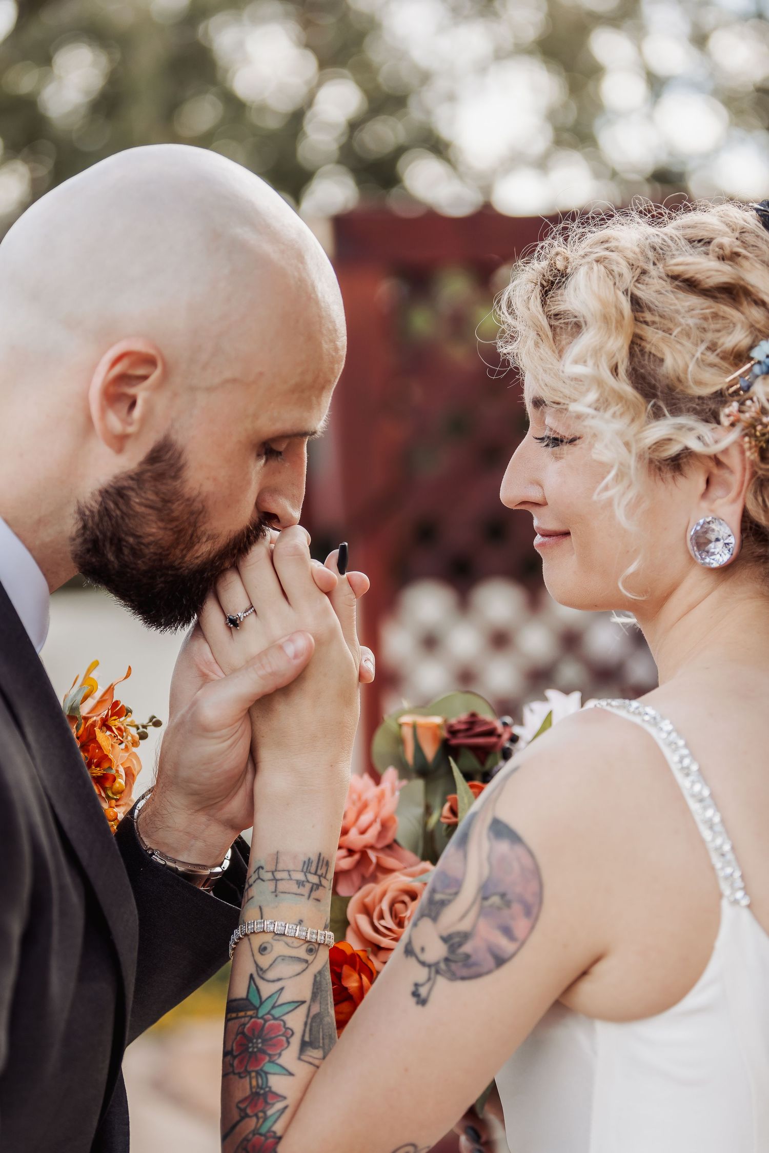 Romantic wedding moment captured outdoors with bride in white dress and groom with beard and tattoos sharing tender gesture.