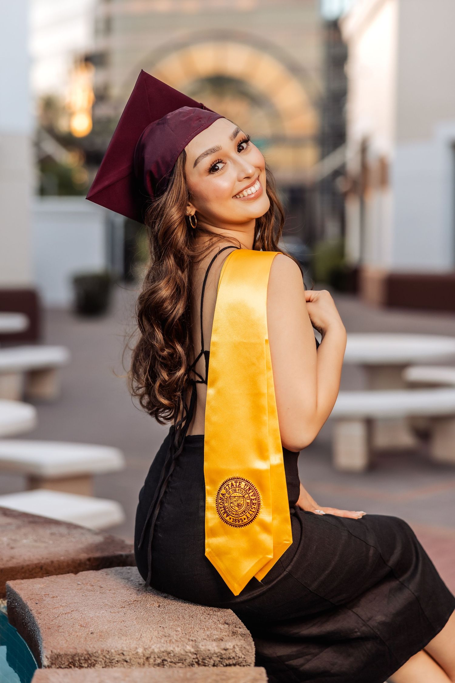Graduate in maroon cap and black dress with gold sash poses outdoors on steps during golden hour celebration.