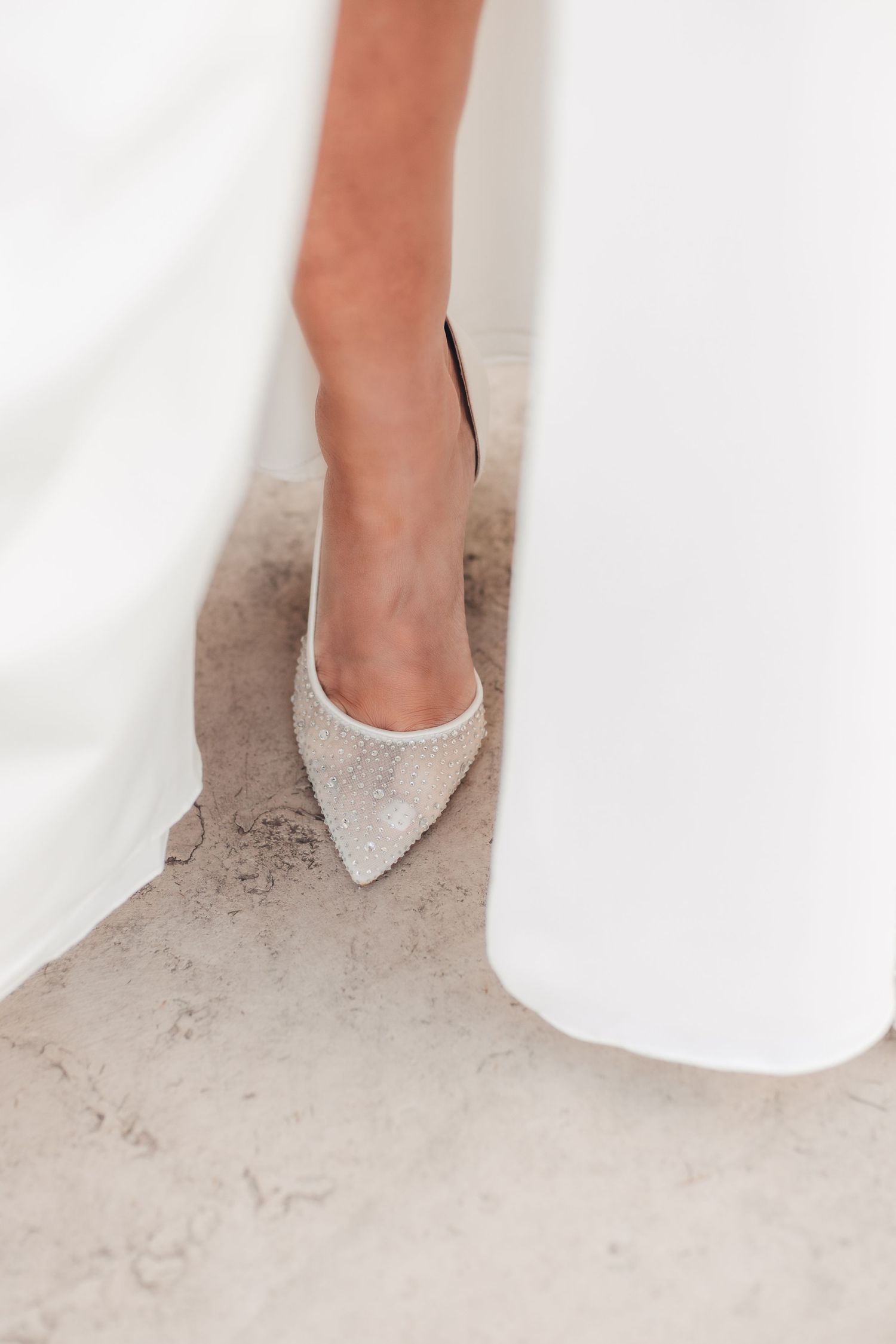 Close-up view of sparkly white pointed-toe bridal shoes peeking out from under a flowing wedding dress.