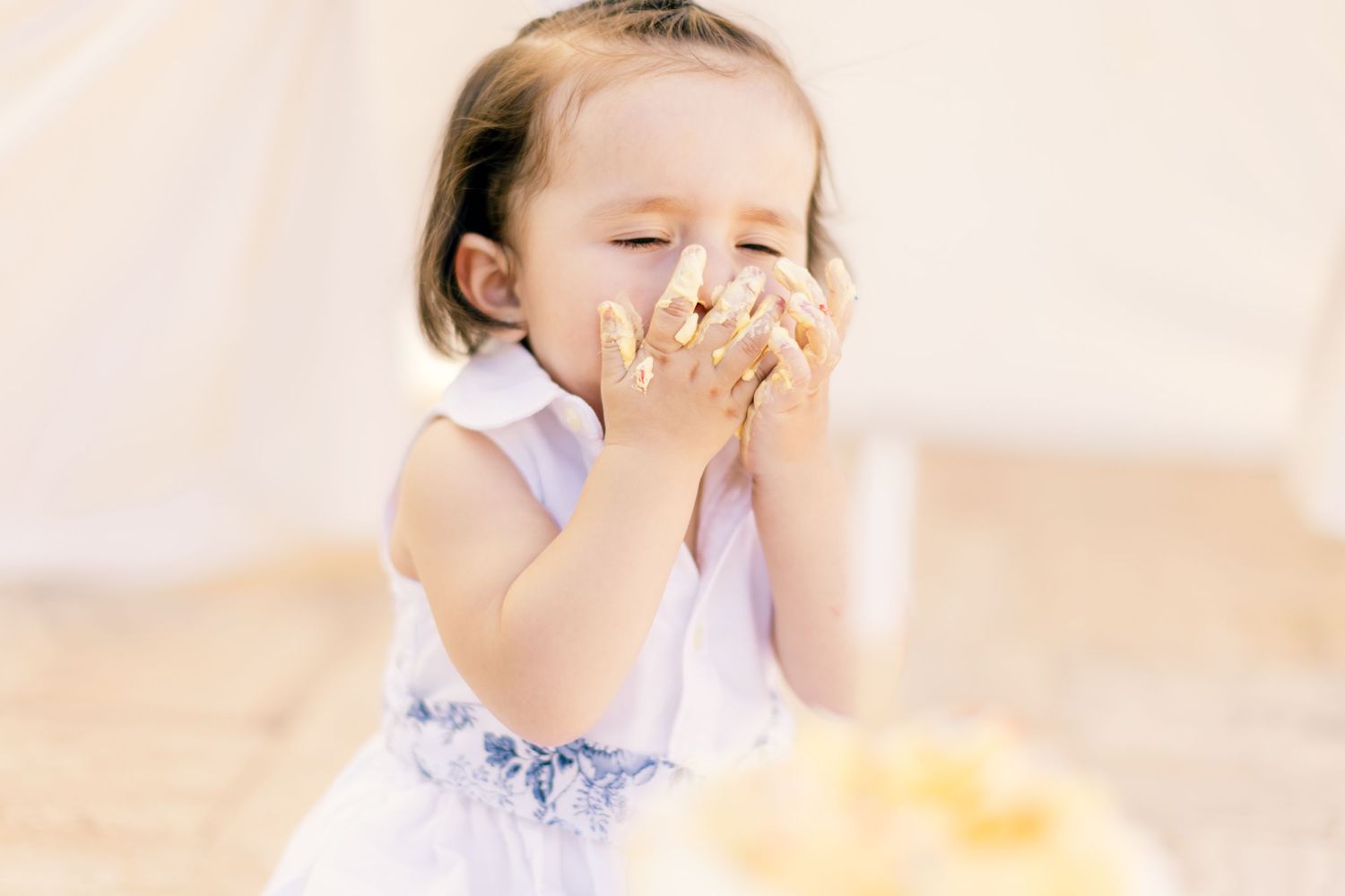 Small child in a white dress covers their mouth while giggling and laughing with bright amusement.