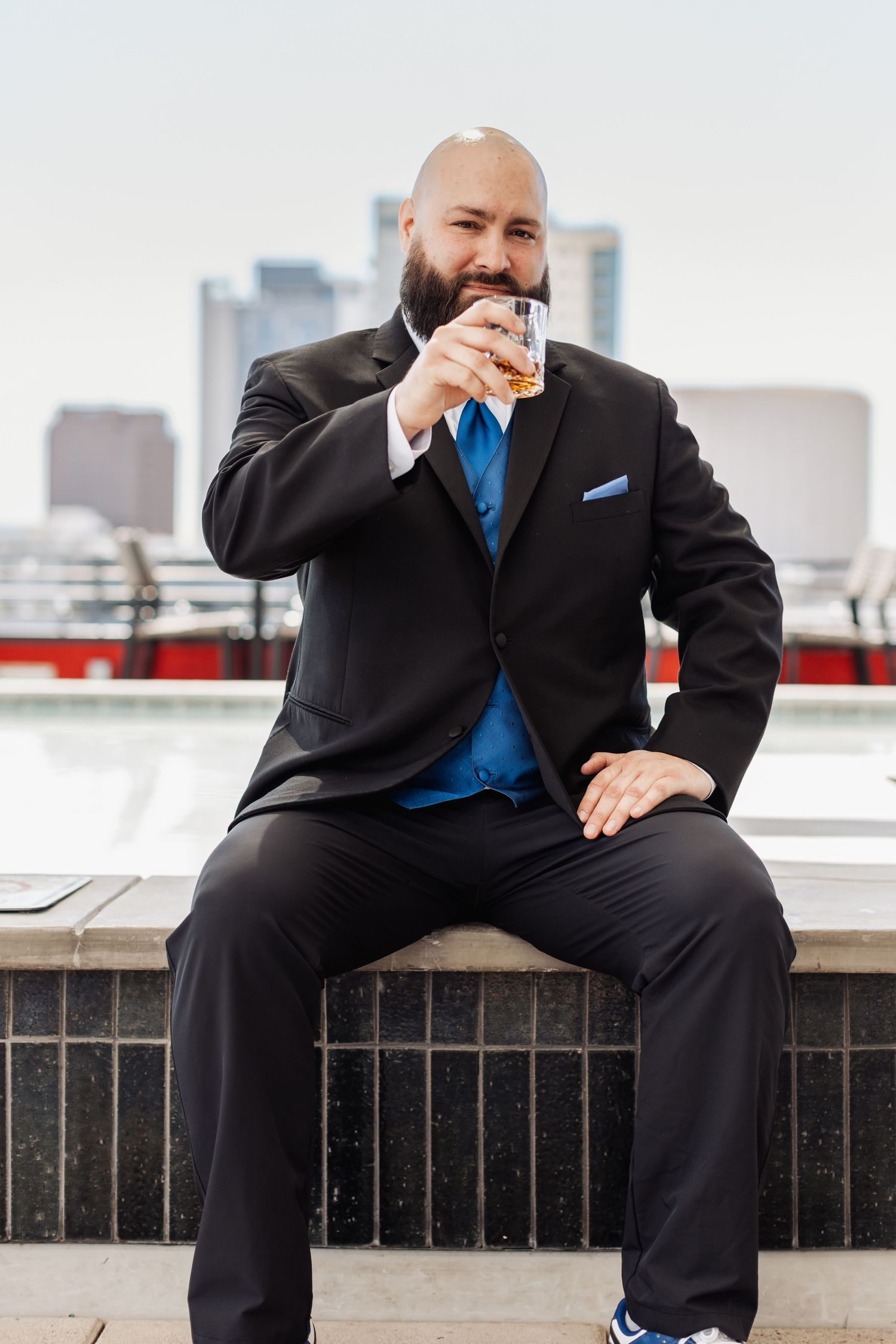 A groom in a black suit and blue tie sits on a ledge with a city skyline in the background.