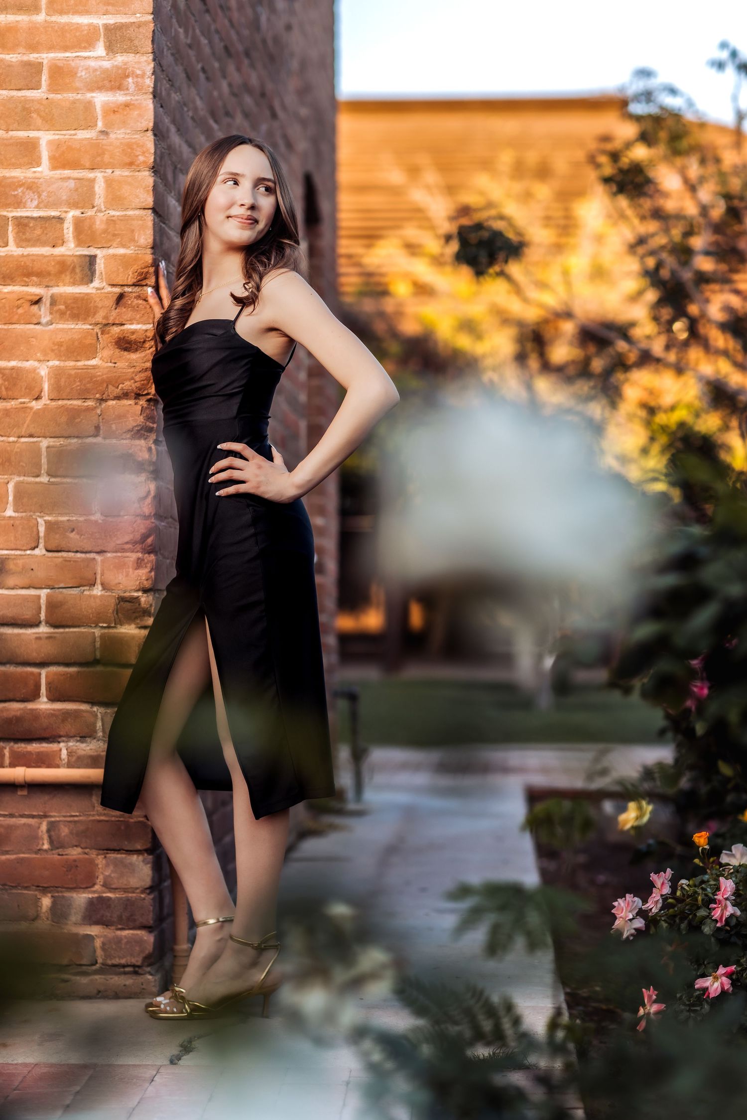 A person in an elegant black dress poses against a brick wall with flowers and foliage in a garden setting at sunset.