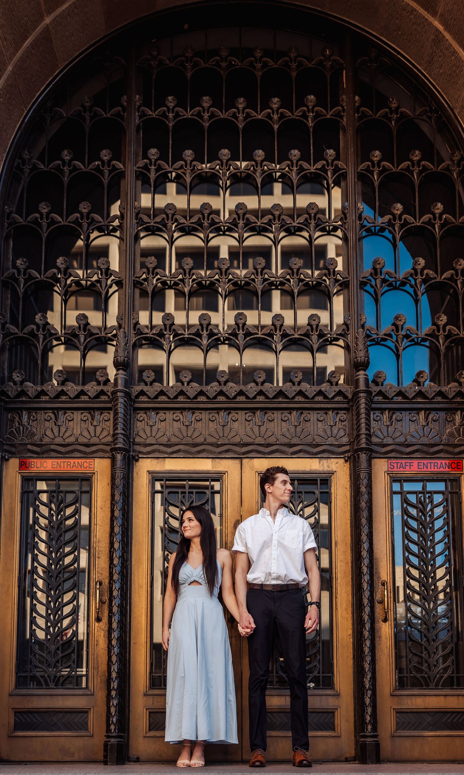 A couple stands in front of ornate vintage doors with intricate black metalwork and gothic architectural details.