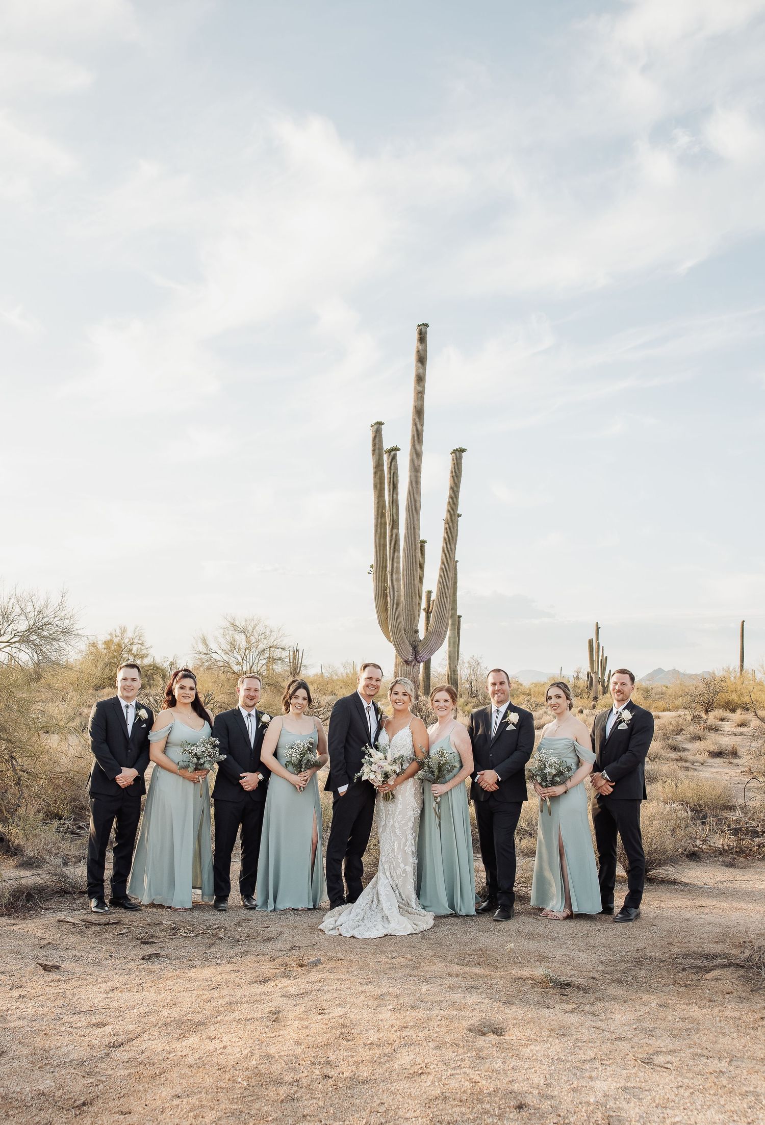 Wedding party poses in front of tall saguaro cacti in desert landscape wearing light blue bridesmaid dresses and black suits.