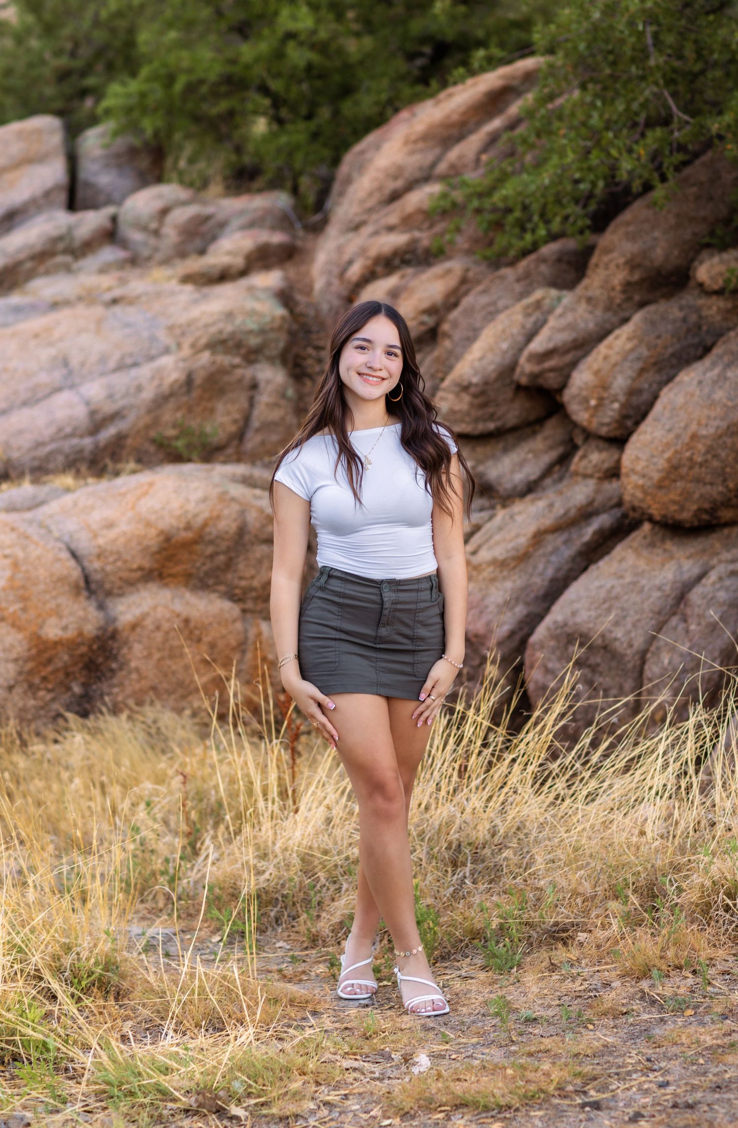 Woman in white t-shirt and dark skirt poses in front of large granite boulders and dried grasses in outdoor setting.