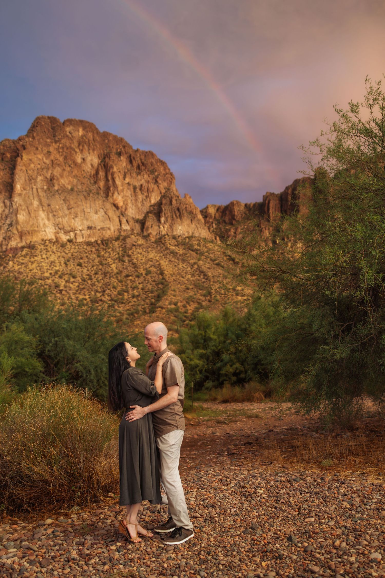 A couple embraces in front of rugged desert mountains with a rainbow arching across the golden evening sky.