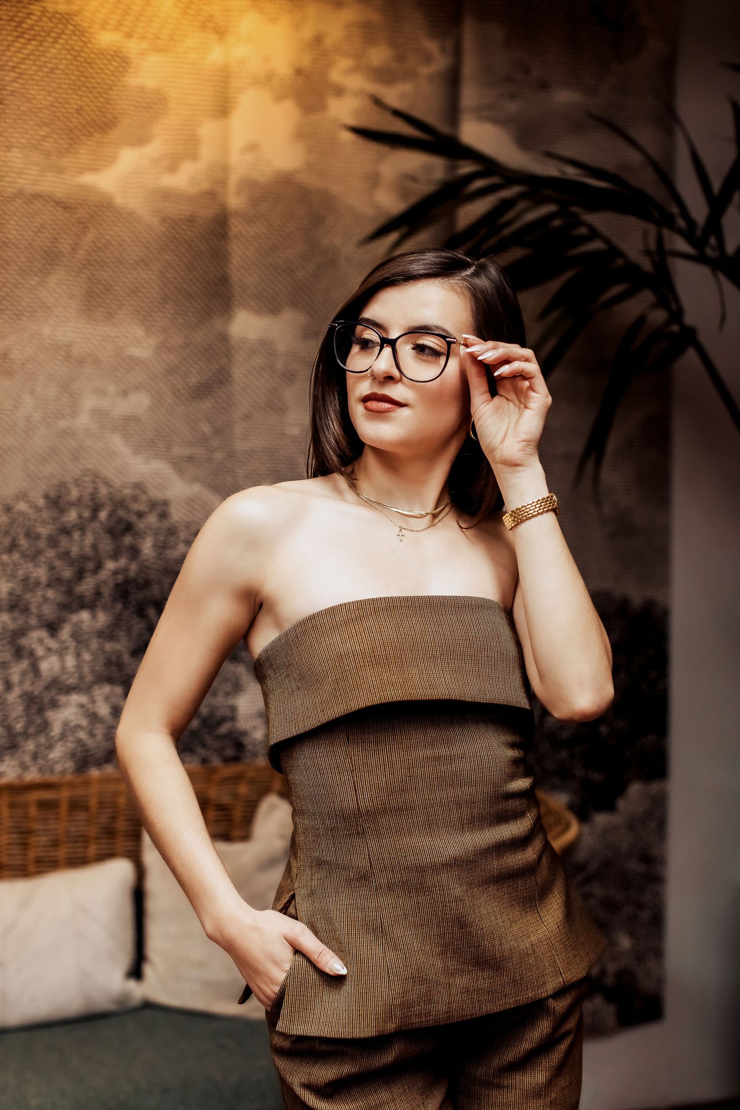 Stylish portrait in brown tones featuring glasses and strapless outfit against tropical foliage background.