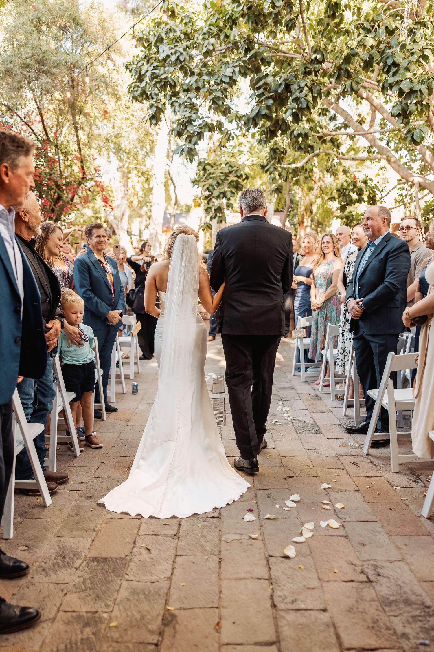 Outdoor wedding ceremony setup with white chairs and flower petals lining the aisle under trees.