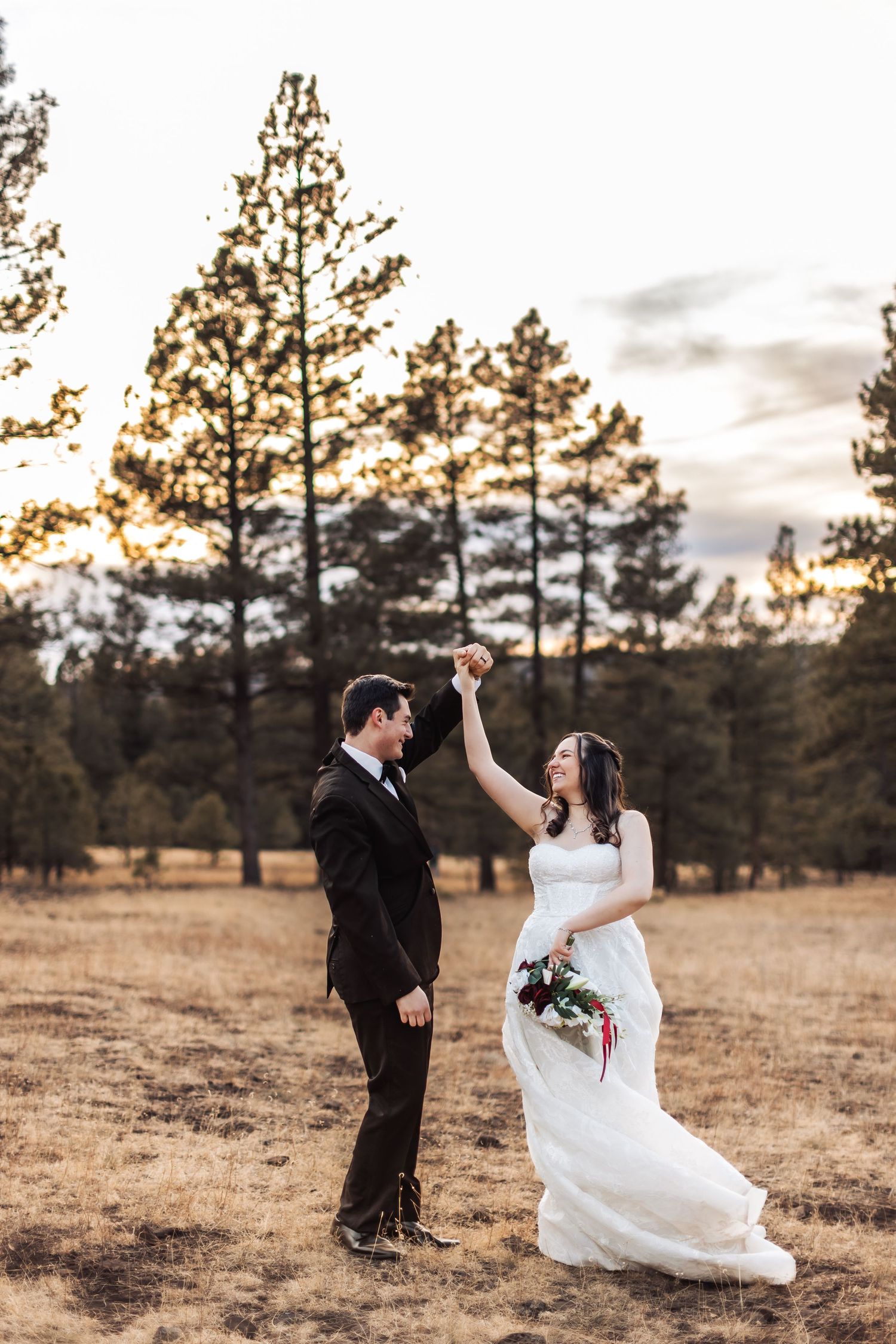 A couple in wedding attire dances together in a golden field at sunset with tall pine trees in the background.