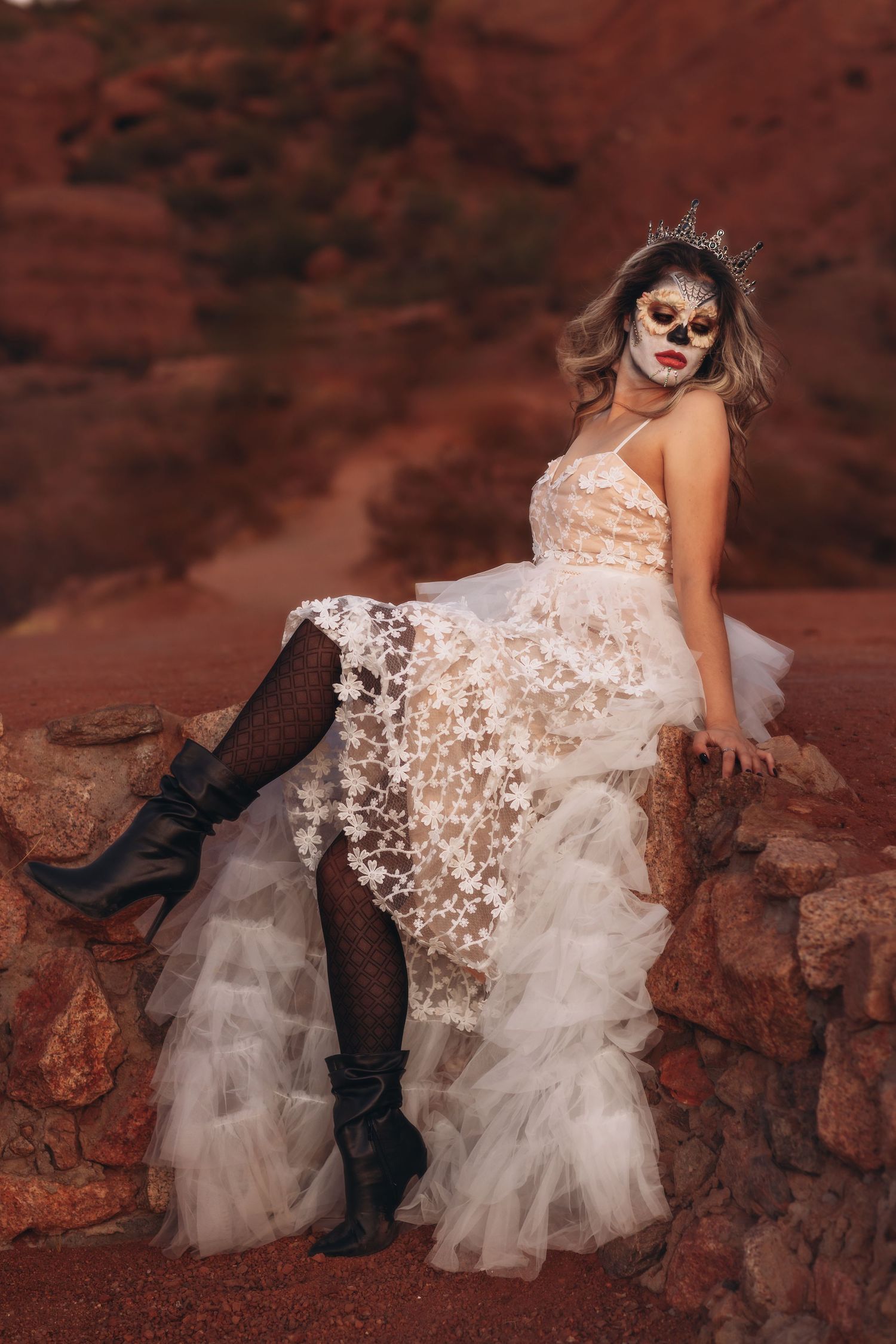 Halloween-themed bridal portrait with skull makeup and lace dress against red rocks showing dark boots and fishnet stockings.