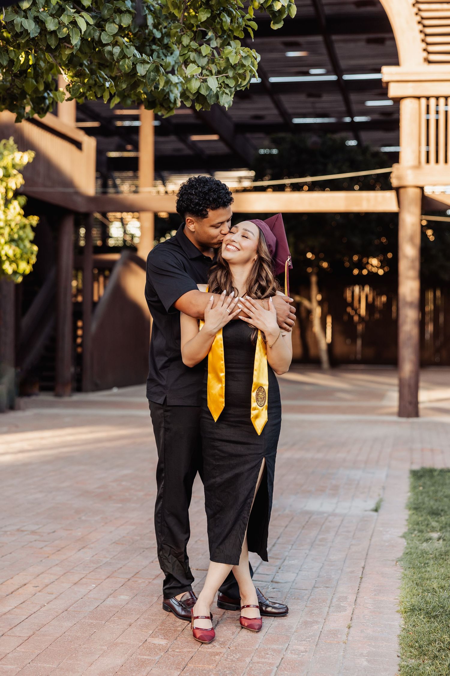 Graduate in cap and gown embraces partner on brick walkway during sunset graduation photoshoot near columns.