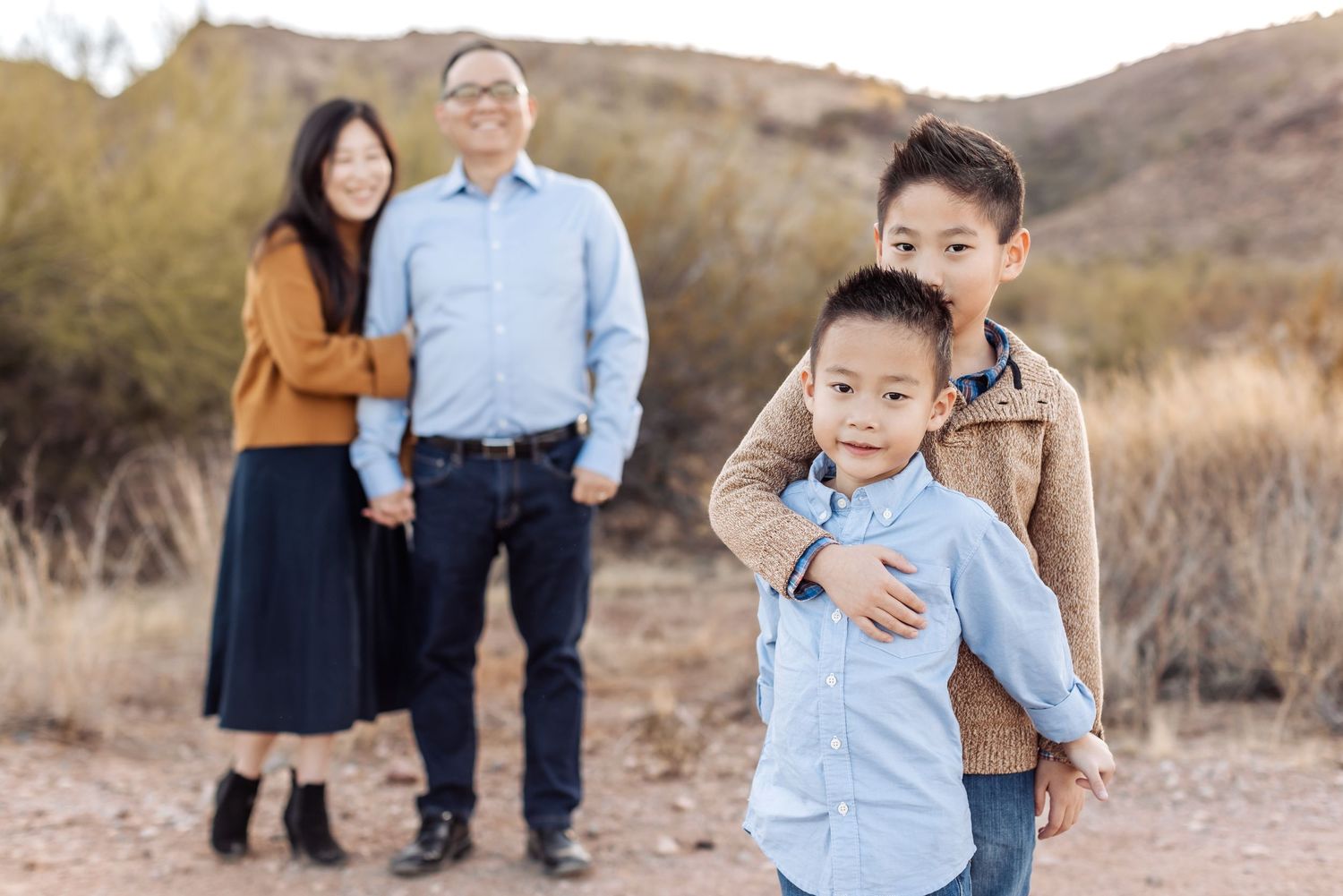 A loving family embraces during an outdoor sunset photoshoot in a desert landscape with brush and mountains.