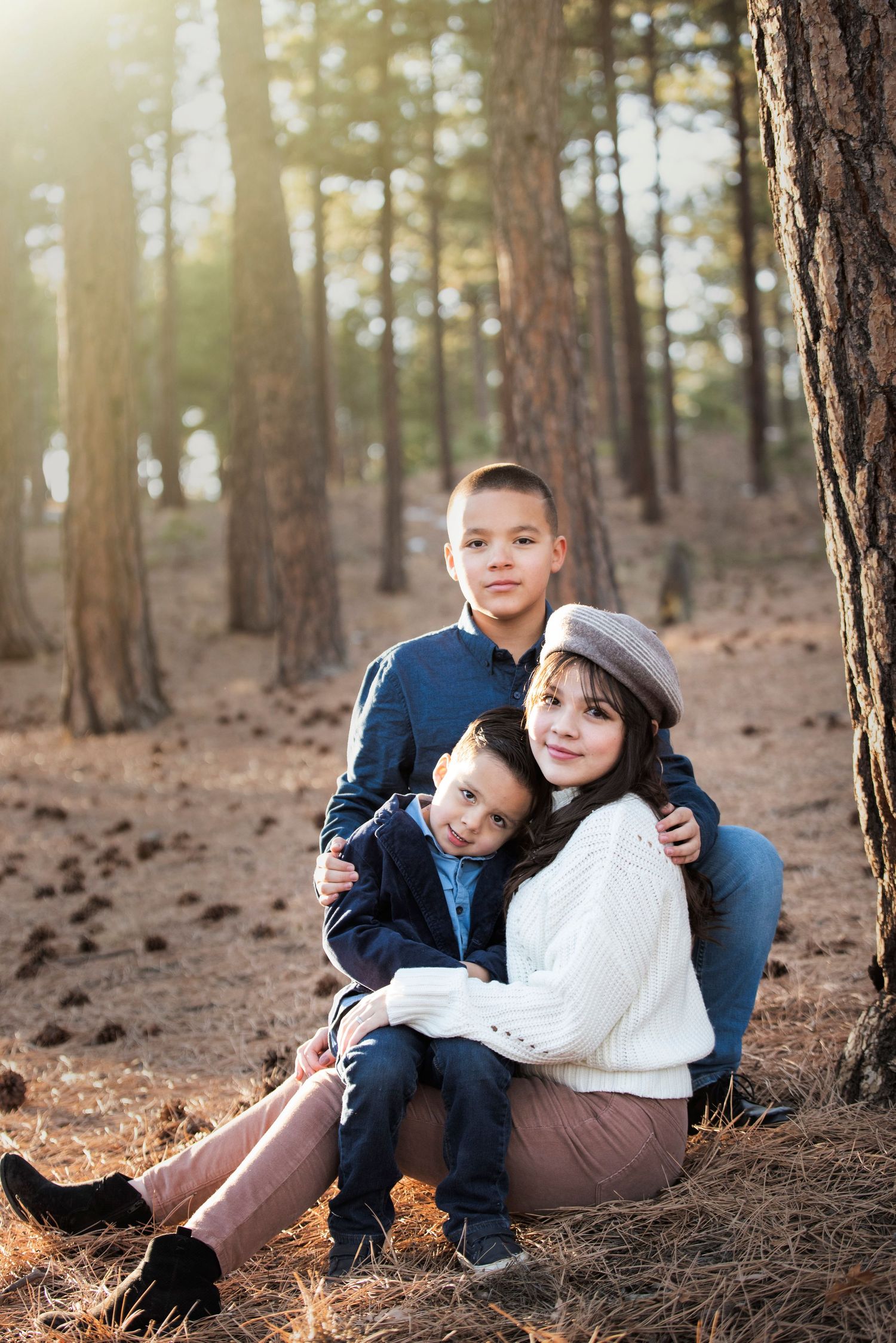 Three siblings sit together on fallen pine needles in a sun-dappled forest during an outdoor photo session.
