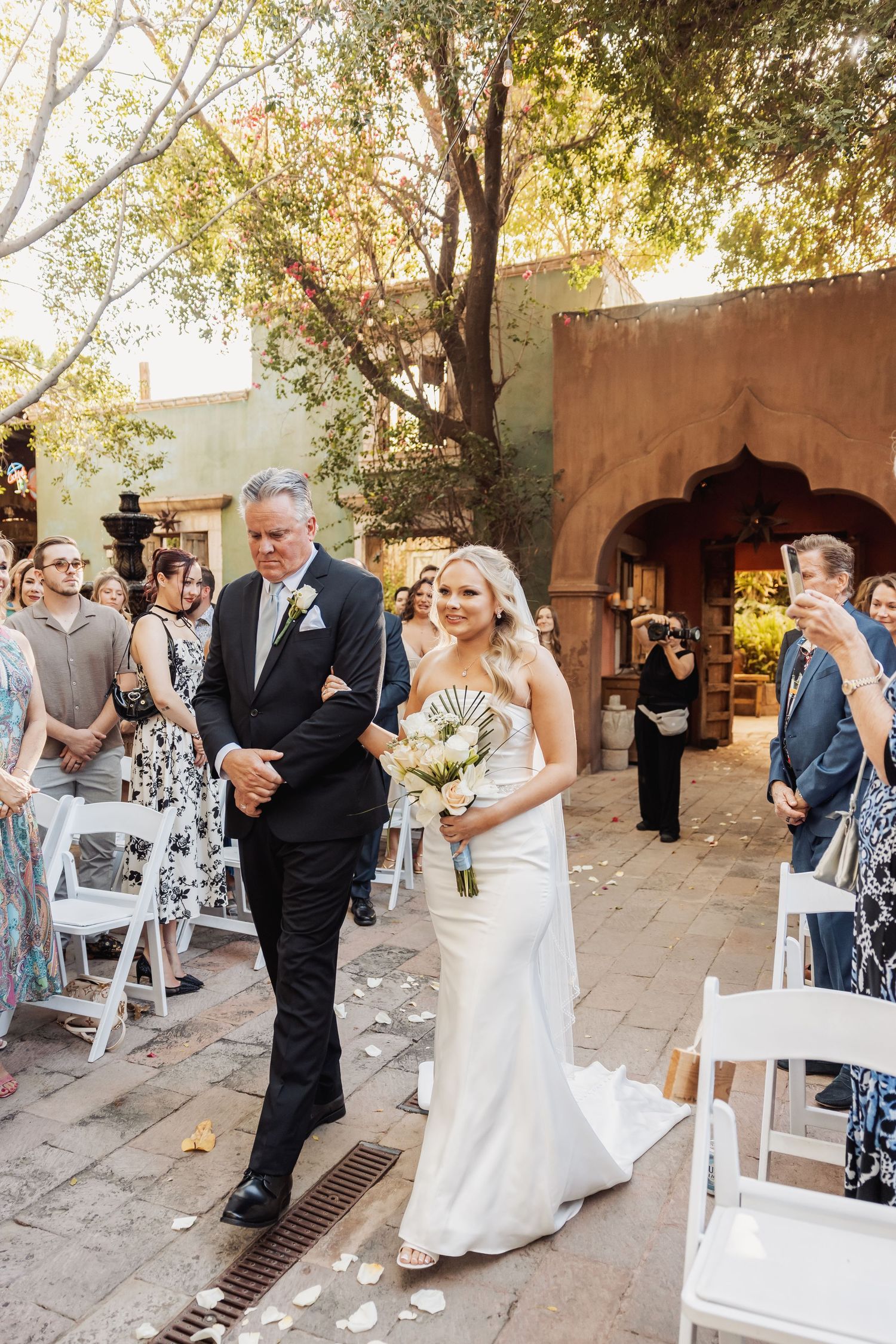 A wedding ceremony takes place in a charming outdoor courtyard with adobe walls and trees providing natural shade.