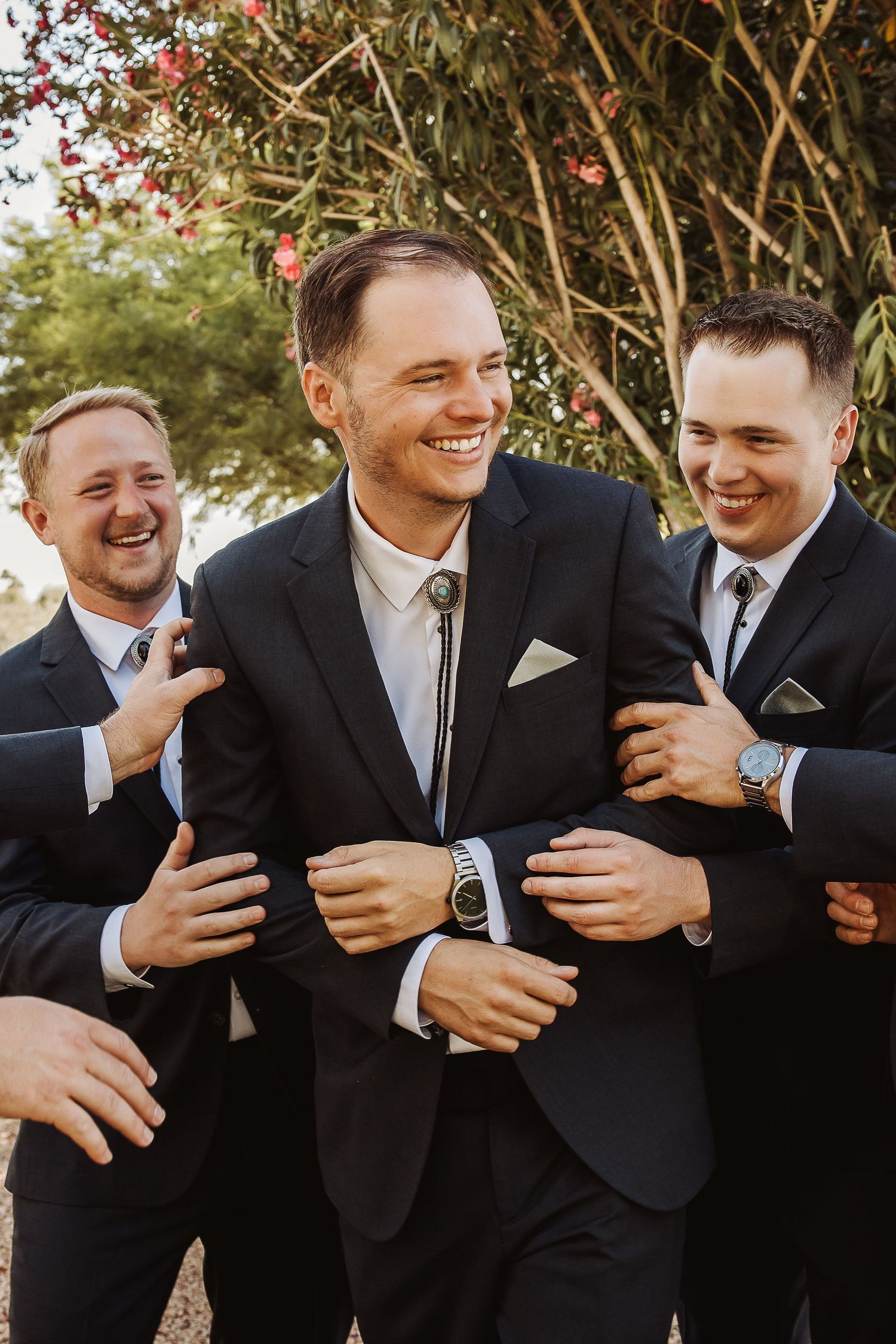 Groomsmen in black suits laughing and embracing in outdoor wedding setting.