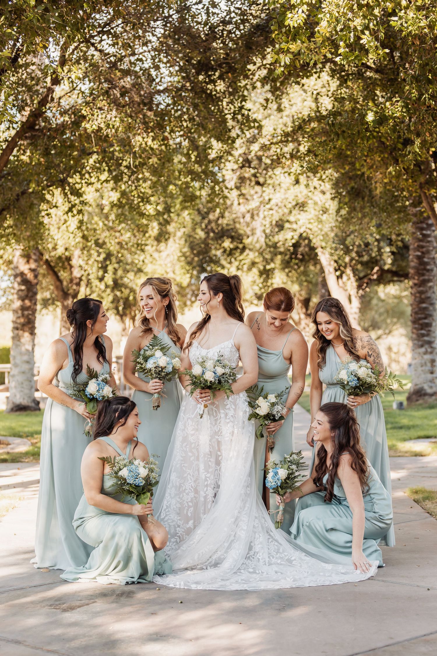 A bridal party in soft sage green dresses poses together outdoors under trees with matching bouquets.