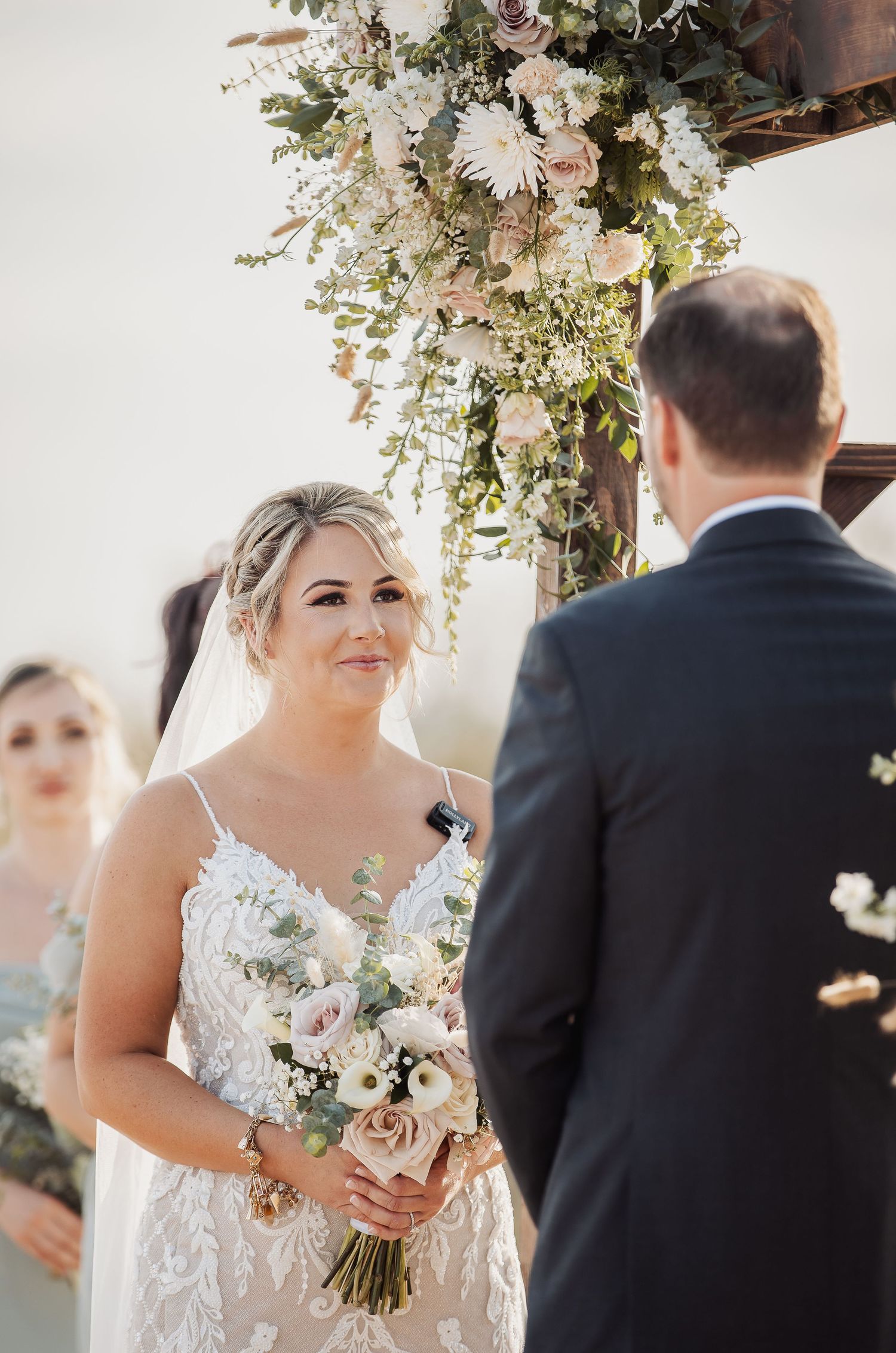 A wedding ceremony under a lush floral arch with white and blush flowers as a couple exchanges vows.